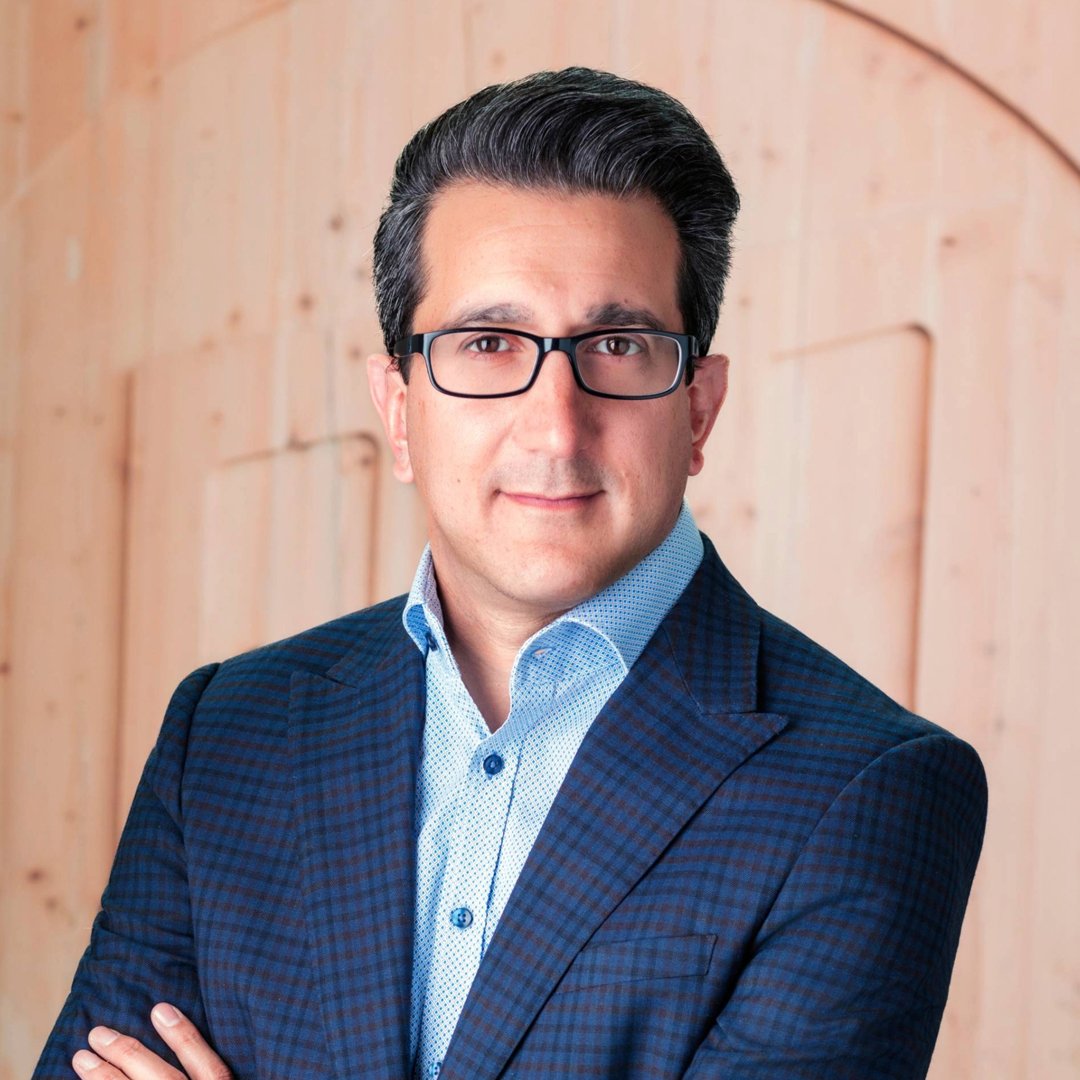 A man with dark hair and glasses wearing a blue checked suit and light blue shirt standing in front of a light wooden background.