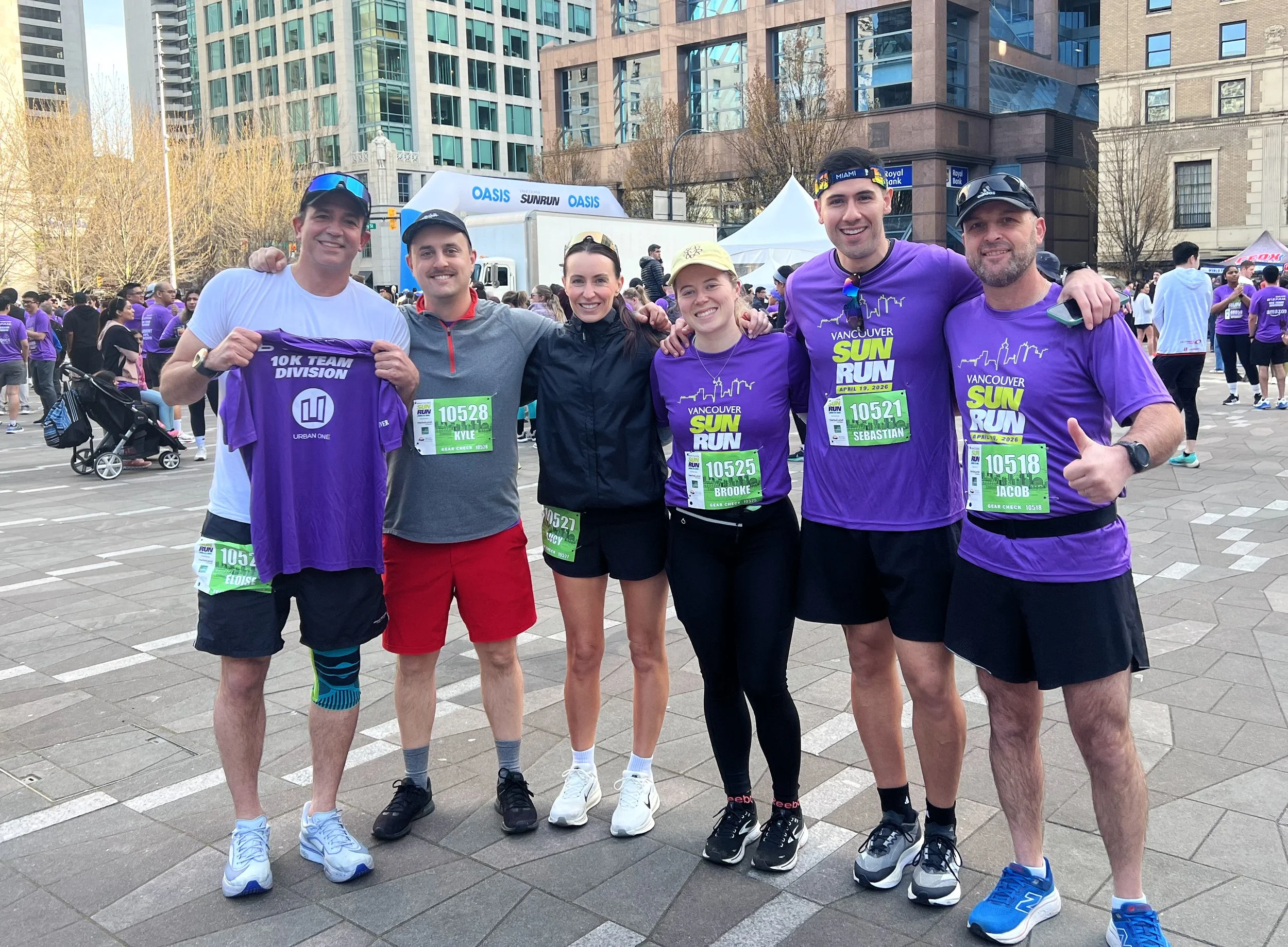 Group of six runners in colorful athletic clothing posing after a race in an urban area, with there race bibs on their chests and a cityscape of tall buildings in the background.