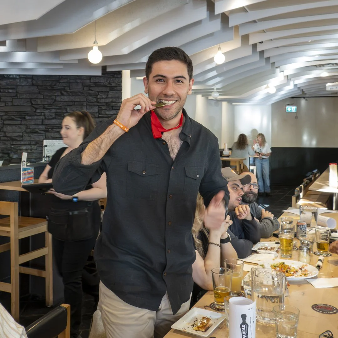 A man smiling and biting into a coin at a restaurant table with several people dining and drinking beer.