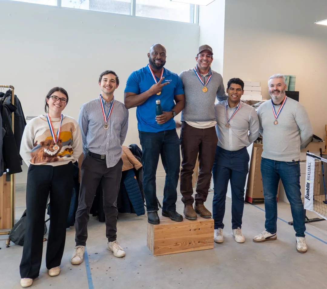 Six people standing in a row with medals, celebrating a victory. One person is on a wooden podium, making a peace sign. Others are smiling, some with hands behind their backs. They are indoors, with a clothing rack, boxes, and a ping pong net visible
