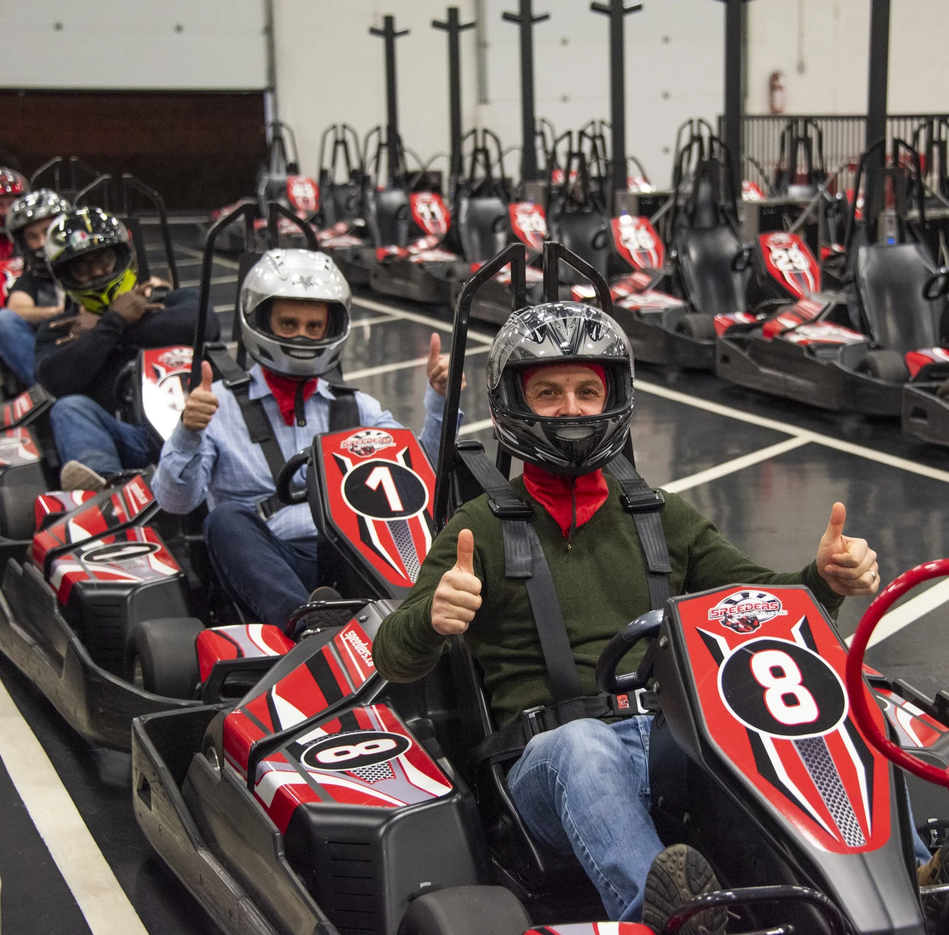 People sitting in go-karts at an indoor racing venue, all wearing helmets and giving thumbs up.