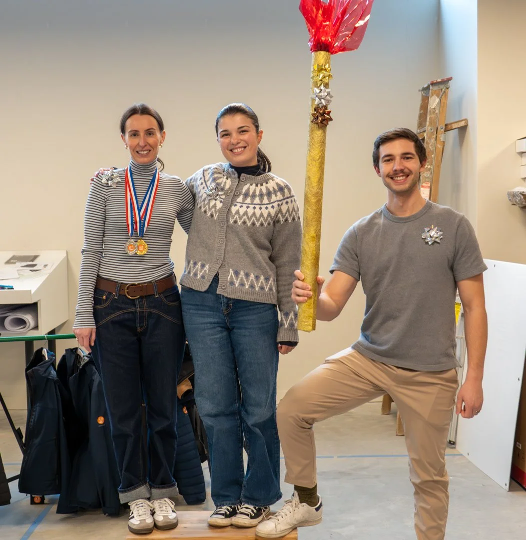 Three people standing indoors, smiling. The woman on the left has medals around her neck and is wearing a striped shirt with a bow on her shoulder. The woman in the middle is wearing a patterned sweater with a bow on her shoulder. The man on the righ