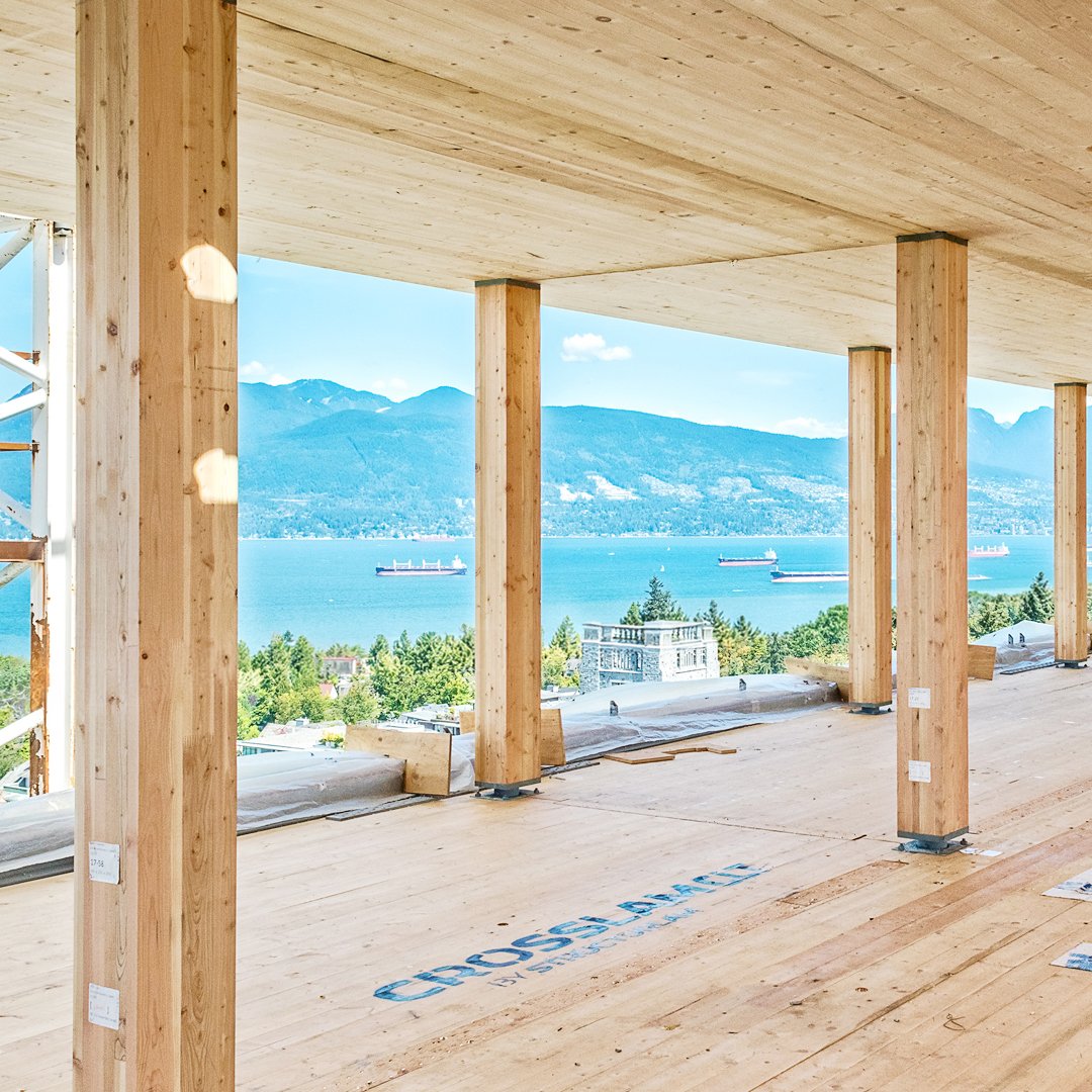 Interior view of a building under construction with large open windows, showing a scenic view of a river, ships, trees, and mountains in the background.