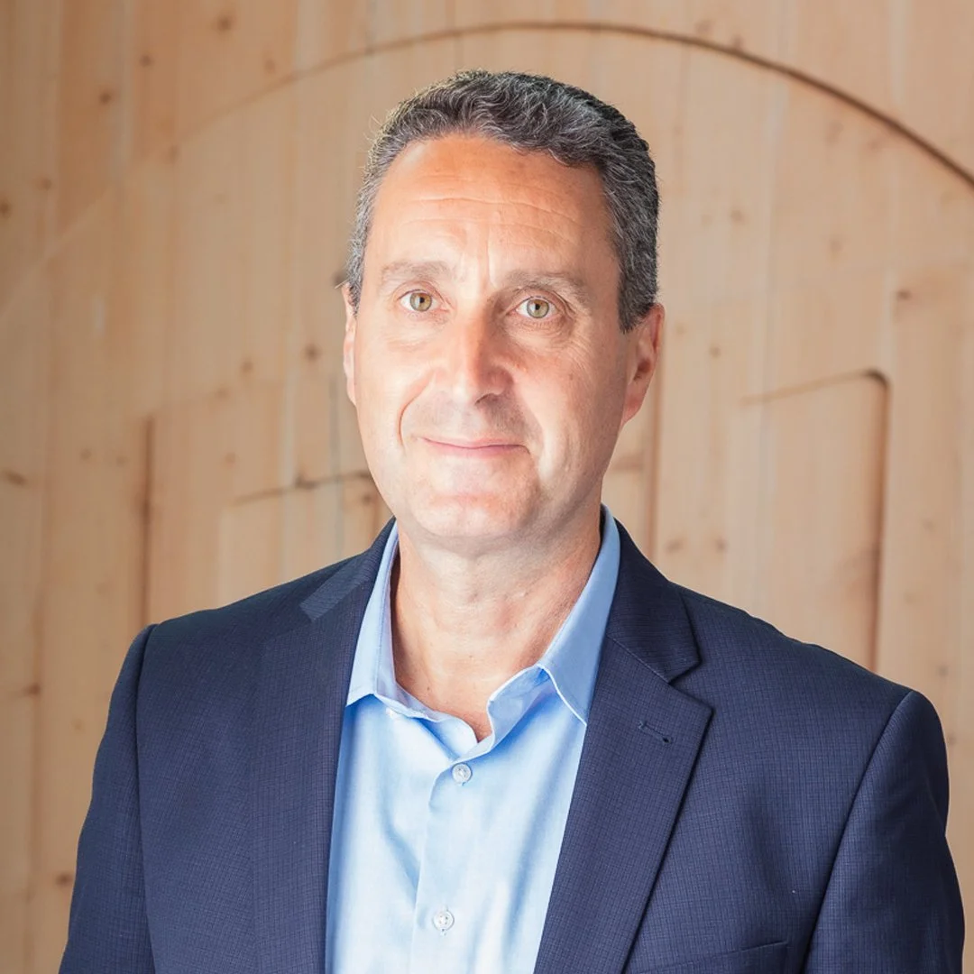 A middle-aged man in a blue suit and light blue shirt posing in front of a wooden background.