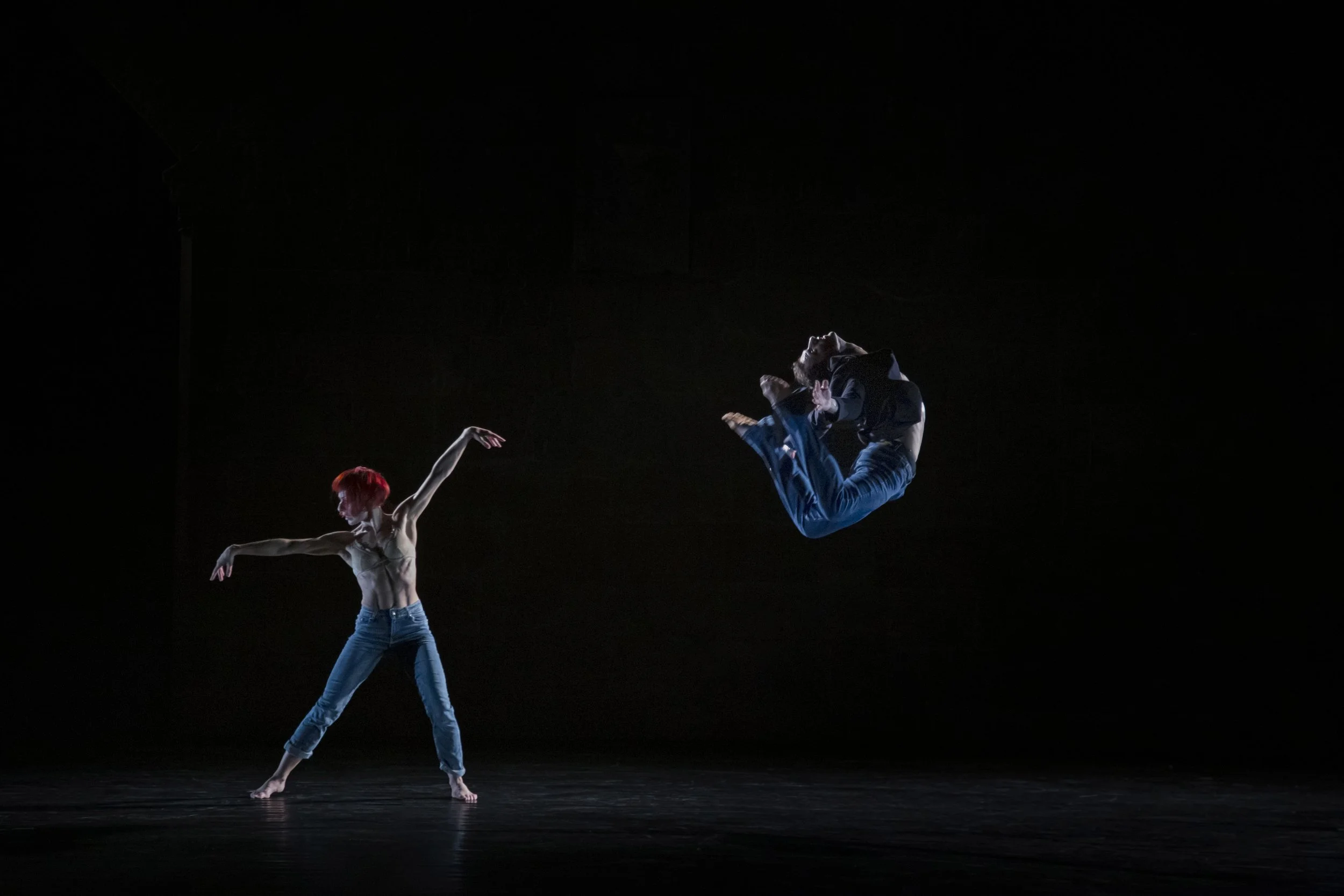 A male dancer performing a backflip while a female dancer stands nearby in a dance performance on a dark stage.