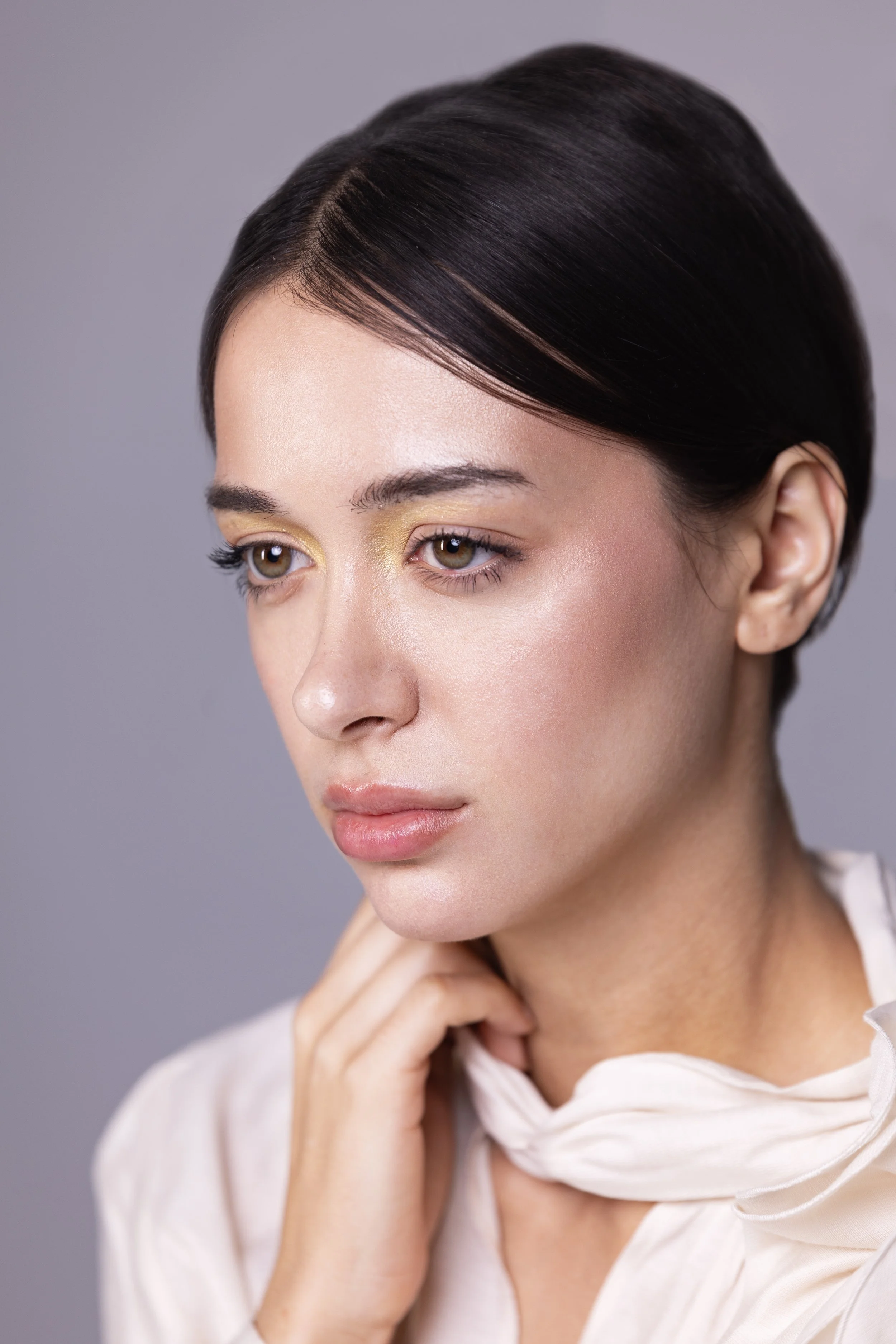 Close-up of a woman with short dark hair, wearing light makeup, touching her neck, against a plain gray background.