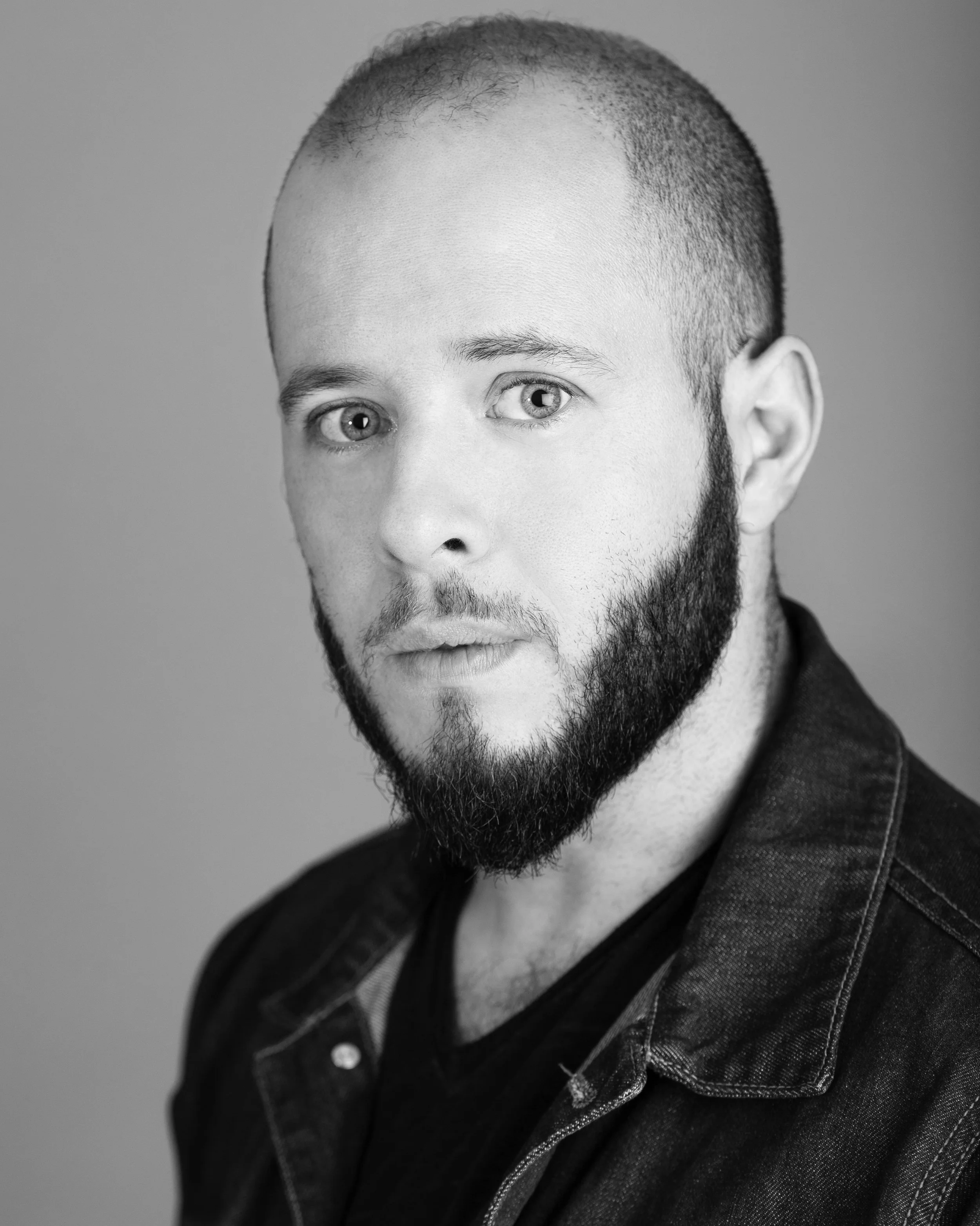 Black and white portrait of a man with a shaved head, beard, and light-colored eyes, wearing a dark jacket.