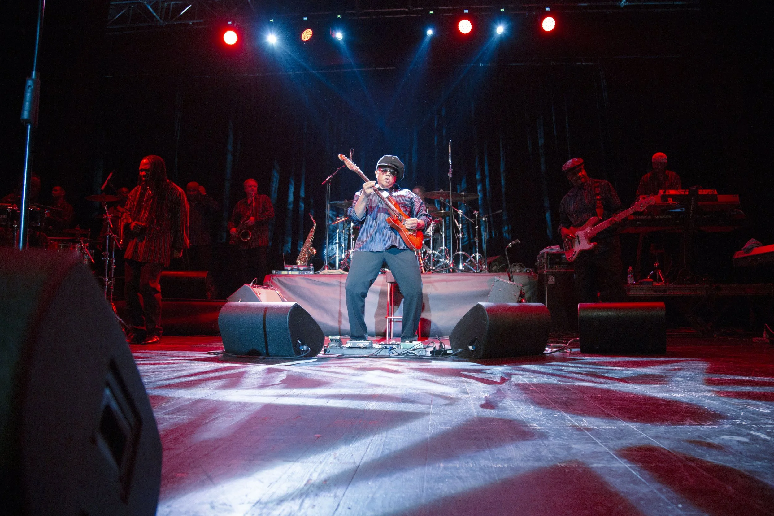 A band performing on stage with a guitarist in front and multiple musicians and instruments in the background, colorful stage lighting illuminating the scene.