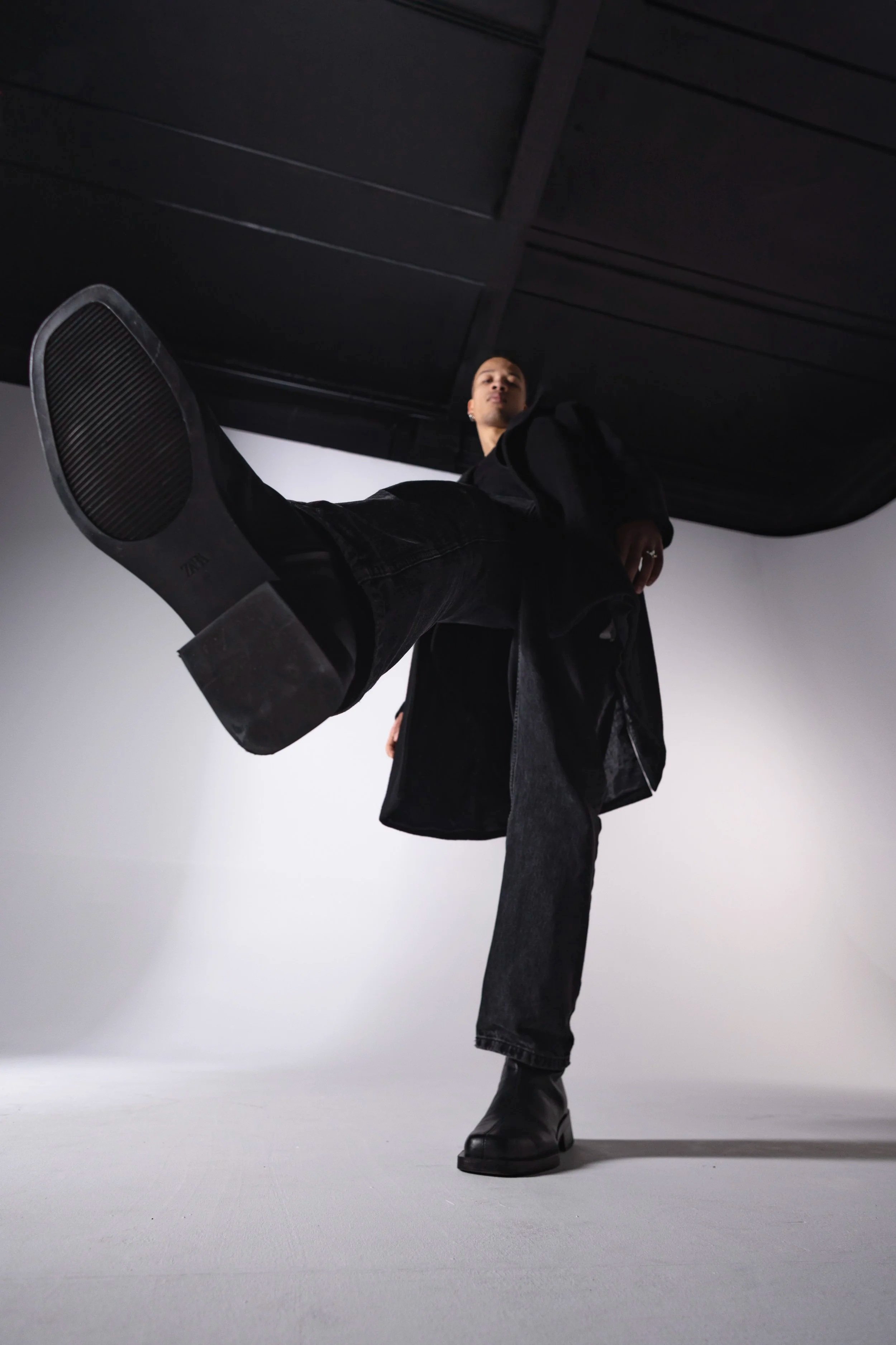 A person wearing black clothing and black boots is standing on a white surface, kicking the camera lens with one foot raised.