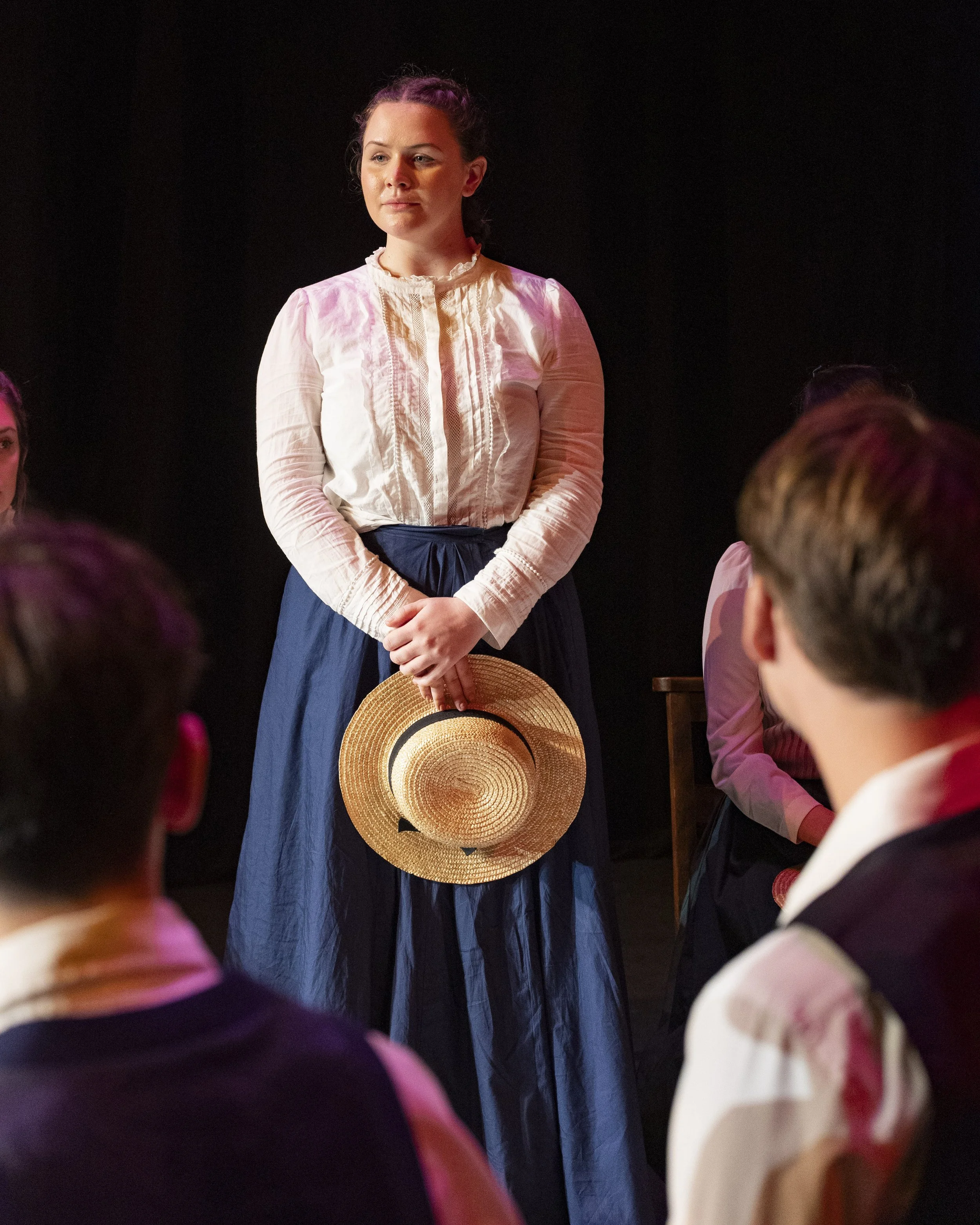 A woman dressed in historical clothing holding a straw hat, standing in front of a group of people in a theatrical or staged setting.