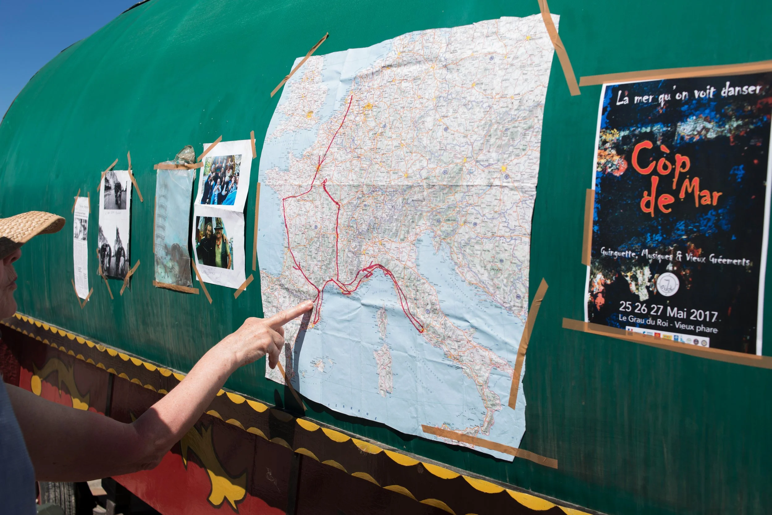 Person pointing at a map of Italy with a red route drawn on it, attached to a green display board with other photographs and a poster for an event in French.