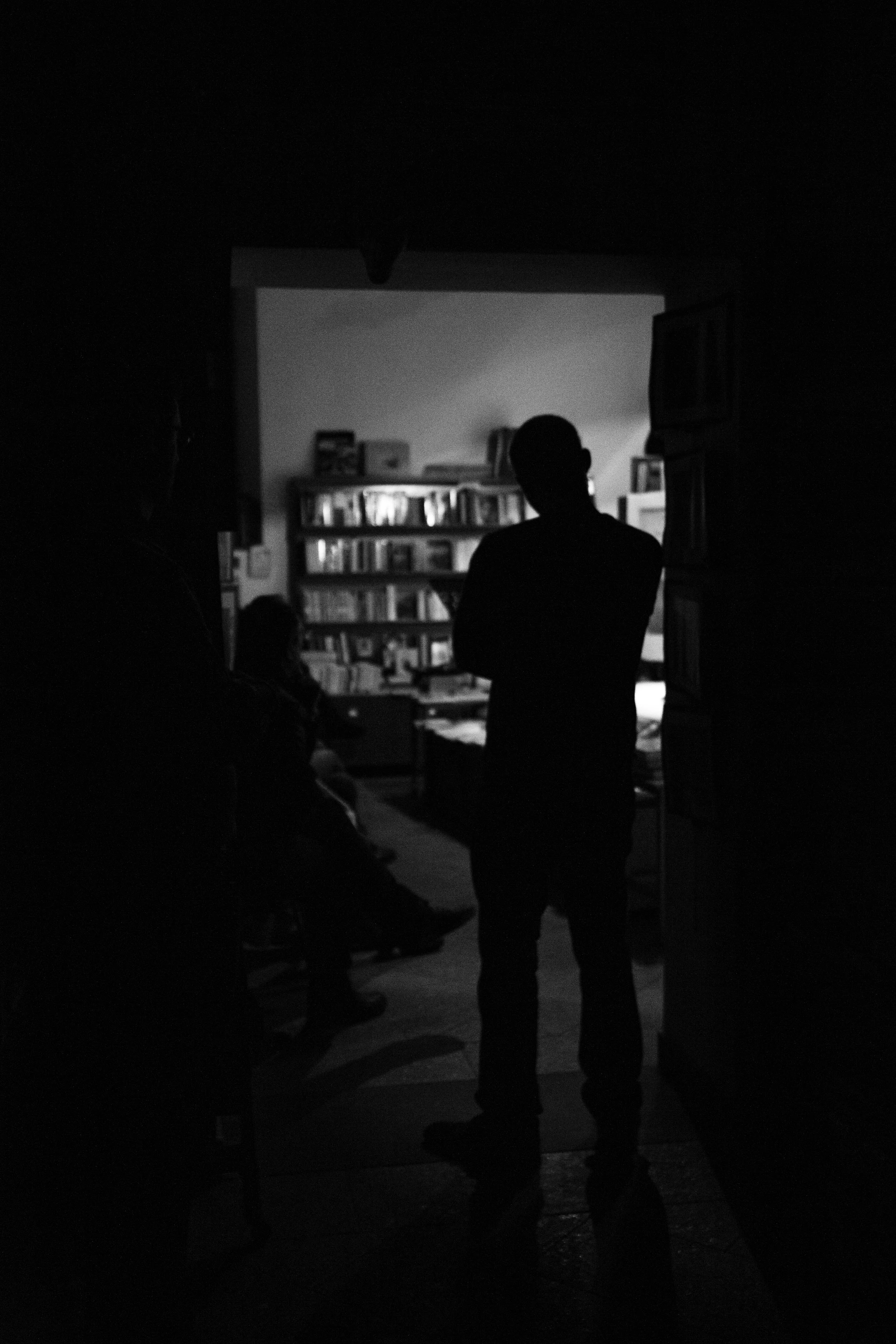 Silhouette of a person standing in a doorway, looking into a well-lit room with bookshelves and people seated inside.