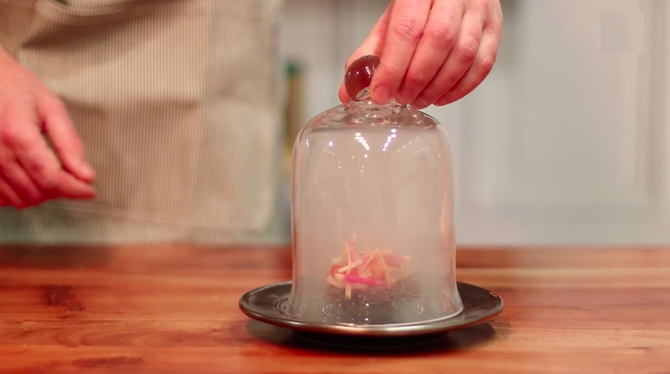 A person using a glass dome cover to reveal a small fine dining. dish with Maine Uni and pickled vegetables on a black plate based on Western Maine.
