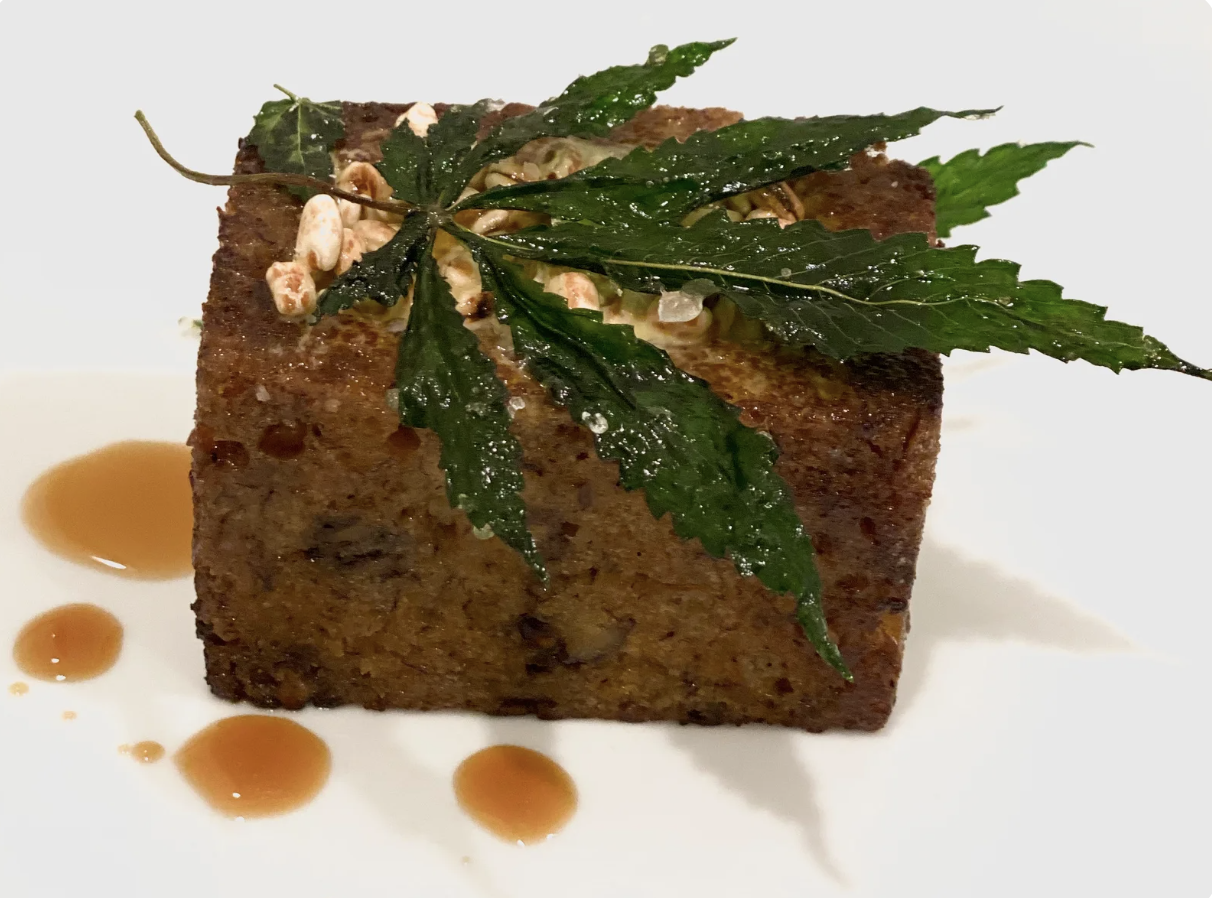 Square piece of banana bread garnished with candied cannabis leaves and topped with nuts, with sauce droplets on the white plate.