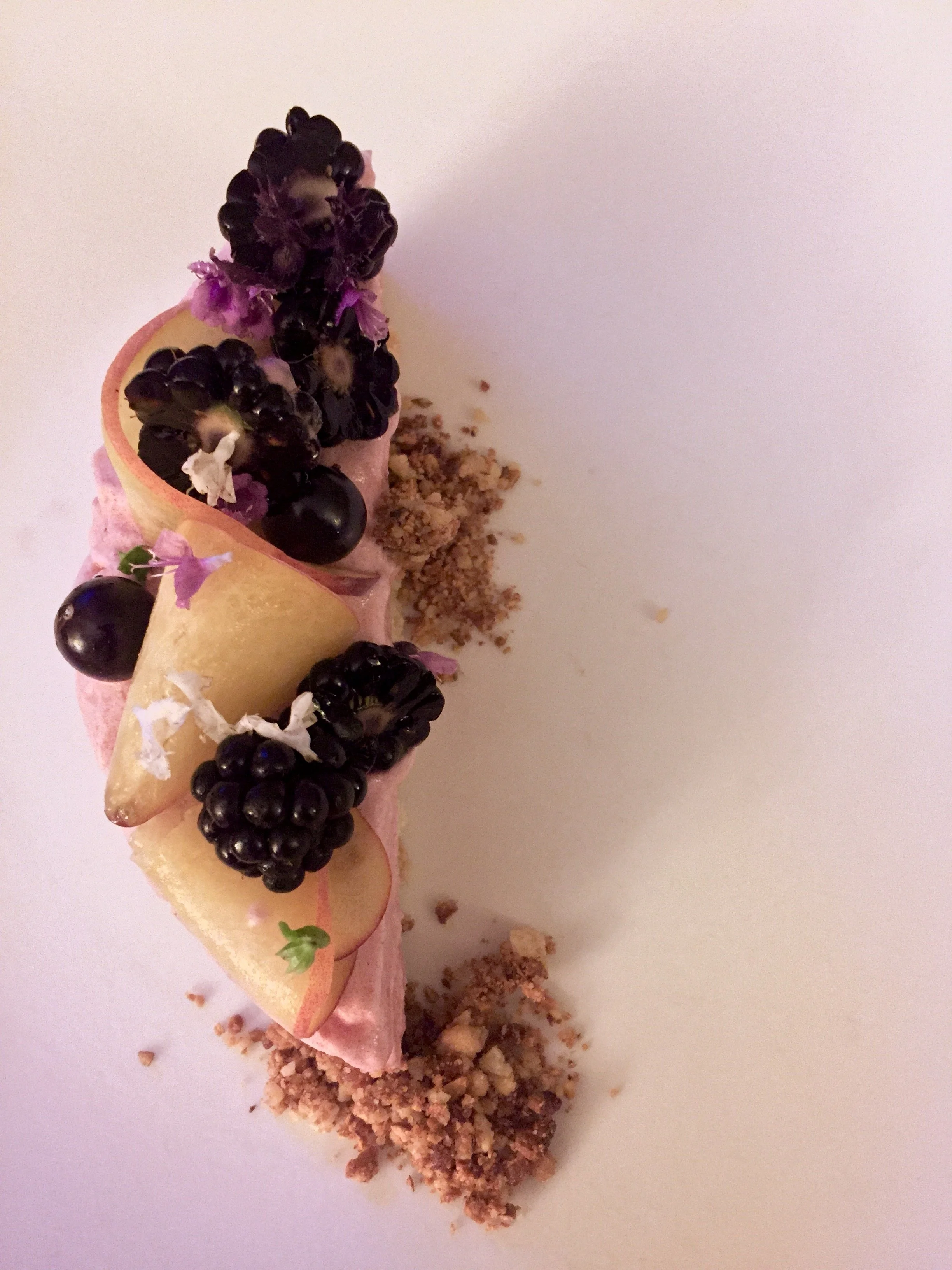 A plated dessert with blackberries, peach slices, edible flowers, pink cream, and crumbled biscuit crumbs on a white plate.