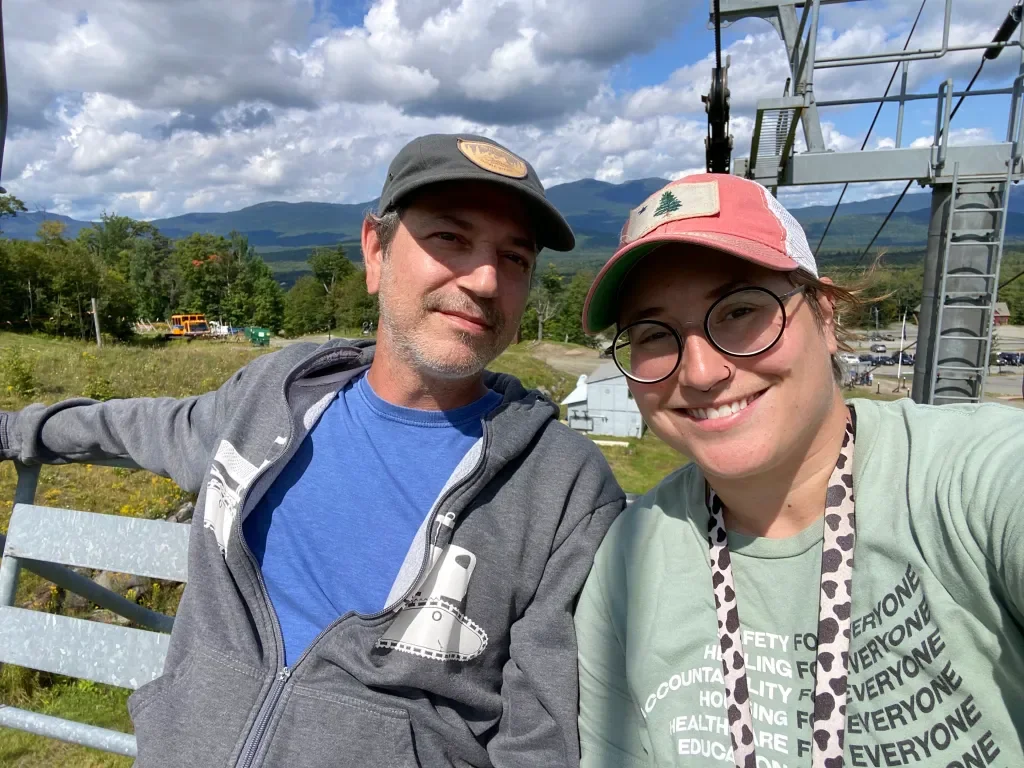 Two people taking a selfie outdoors on a cloudy day with Maine mountains and a ski lift in the background.