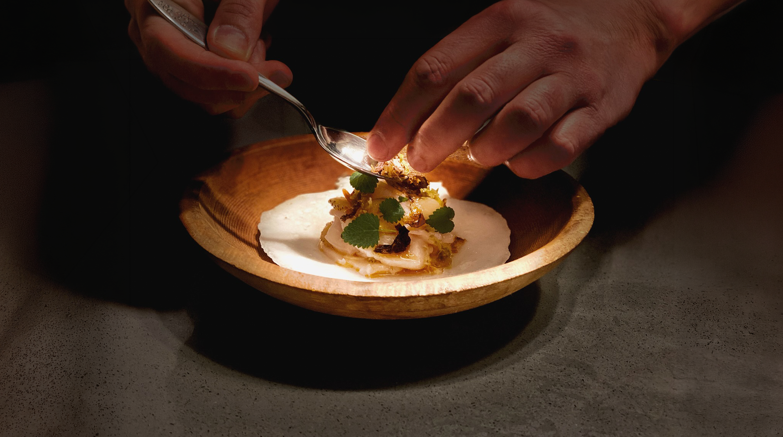 Chef garnishing a fine dining dish of Maine sea scallops in the shell. with herbs and crispy toppings on a wooden plate.
