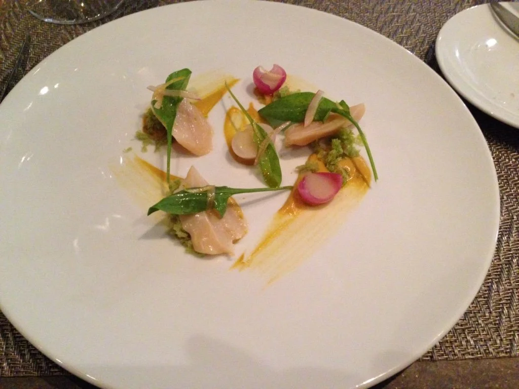 A white plate with a gourmet appetizer of sliced radishes, green leaves, and small bites of fish or seafood, garnished with a light sauce and herbs.