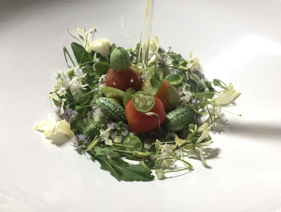 A floral arrangement made of toy vegetables, including watermelons, a tomato, and a gherkin, surrounded by small flowers and green leaves.