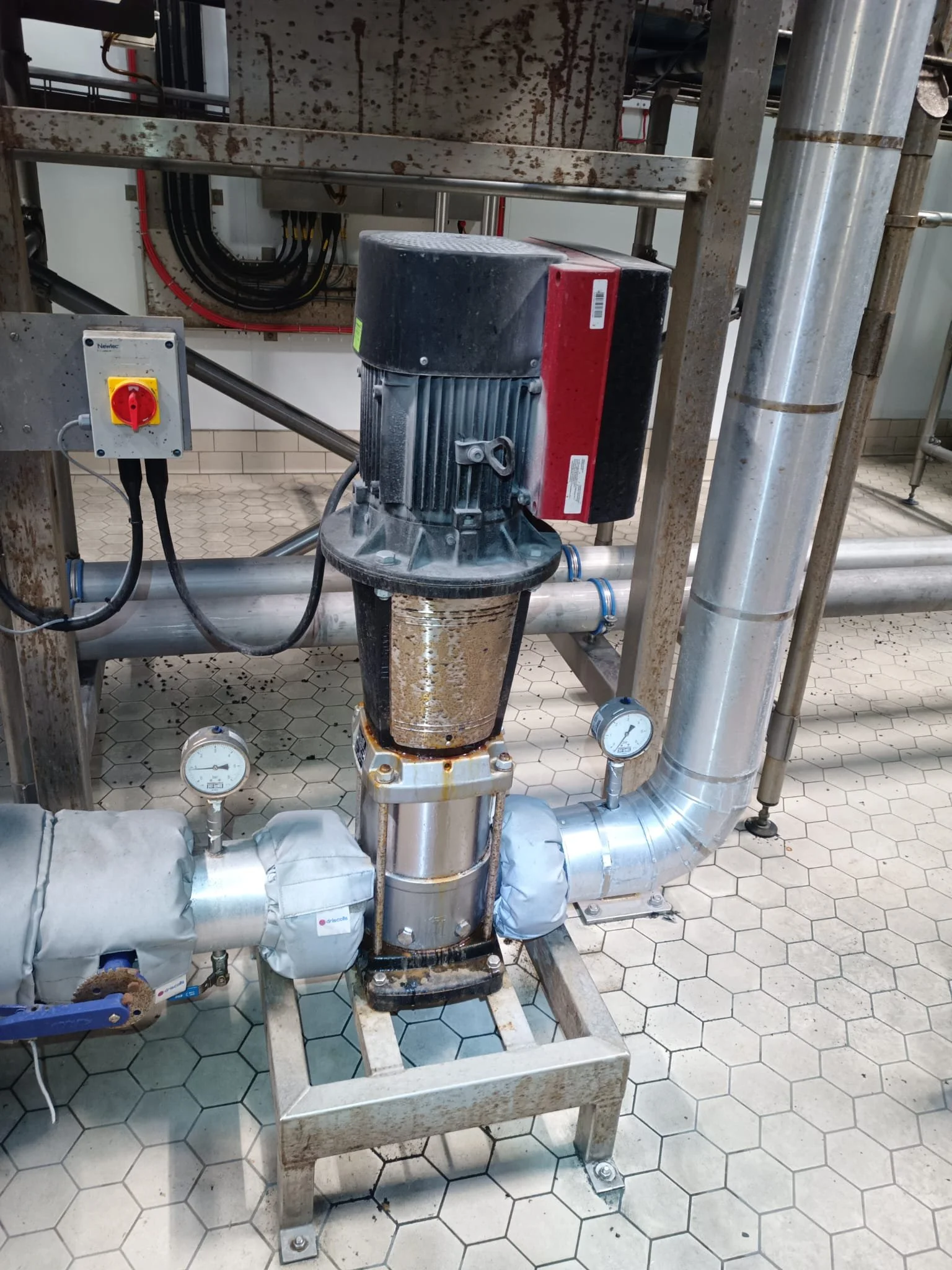 Industrial pump connected to pipes with gauges and a rusty metal frame.