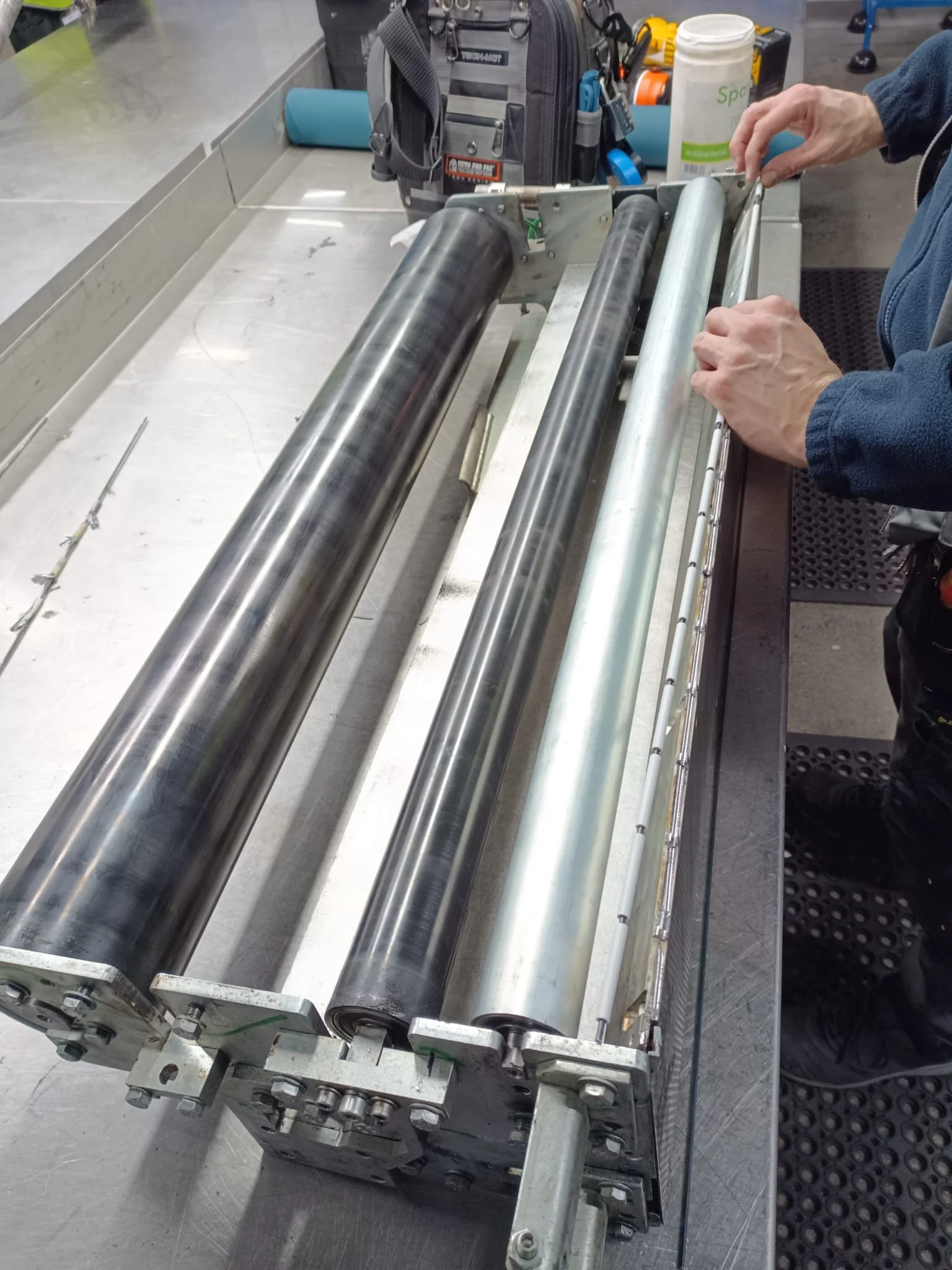 Close-up of an industrial roller press machine on a work table, with a person's hands adjusting it. The background includes a toolbox and cleaning supplies.