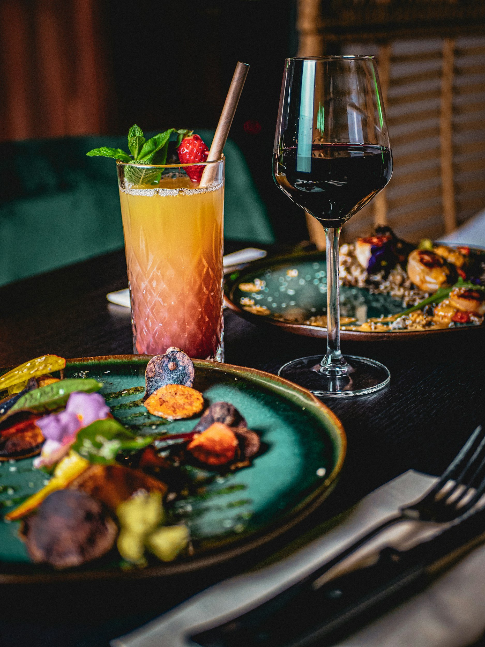 A dining table set with a colorful cocktail garnished with a strawberry and mint, a glass of red wine, and plates with food.