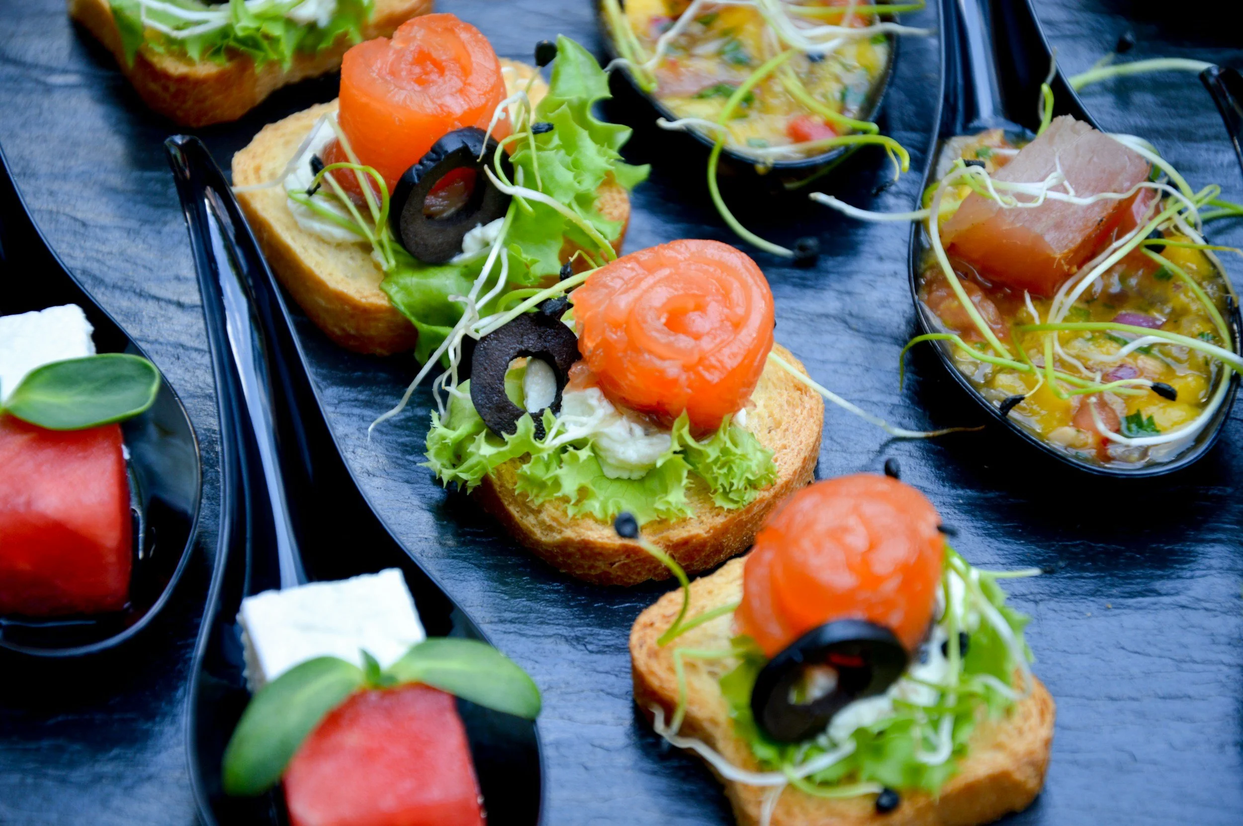 Assorted appetizers on black serving platters, including bread topped with greens, smoked salmon, black olives, and creamy spread, alongside small bowls of colorful soup with vegetables and sprouts.