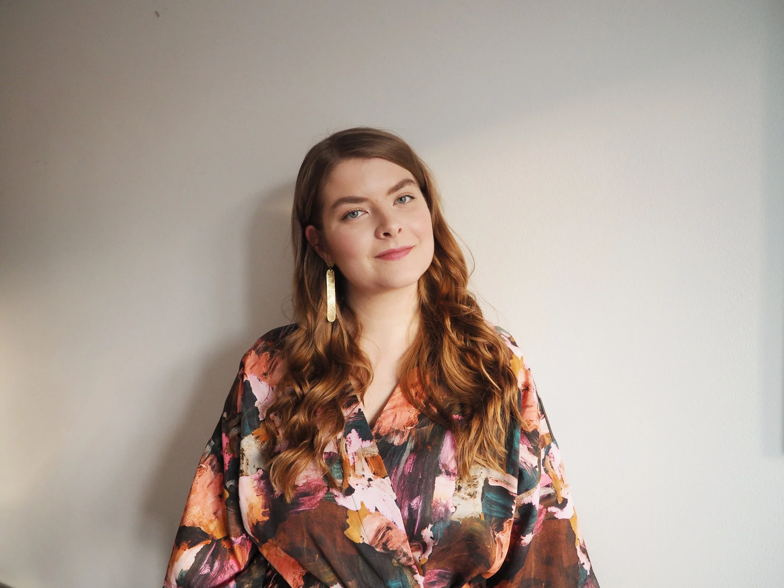 A young woman with long, wavy auburn hair and light skin poses against a plain, light-colored wall, wearing a colorful floral patterned blouse and long gold earrings.