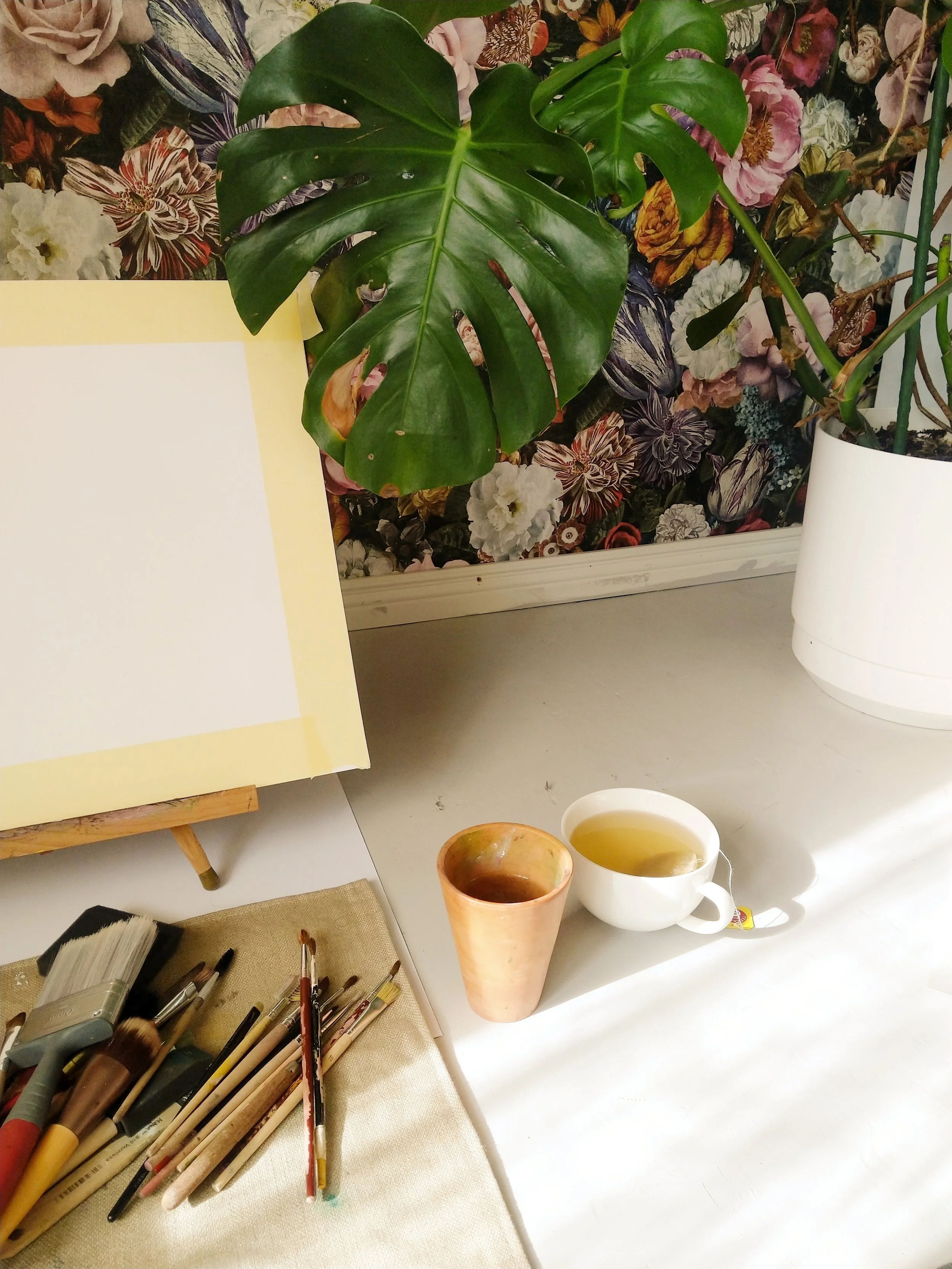 A table with two cups of tea or coffee, one in a white mug and the other in a ceramic cup, a collection of paint brushes on a beige cloth, a large green monstera plant with split leaves, and a background of floral patterned wallpaper.