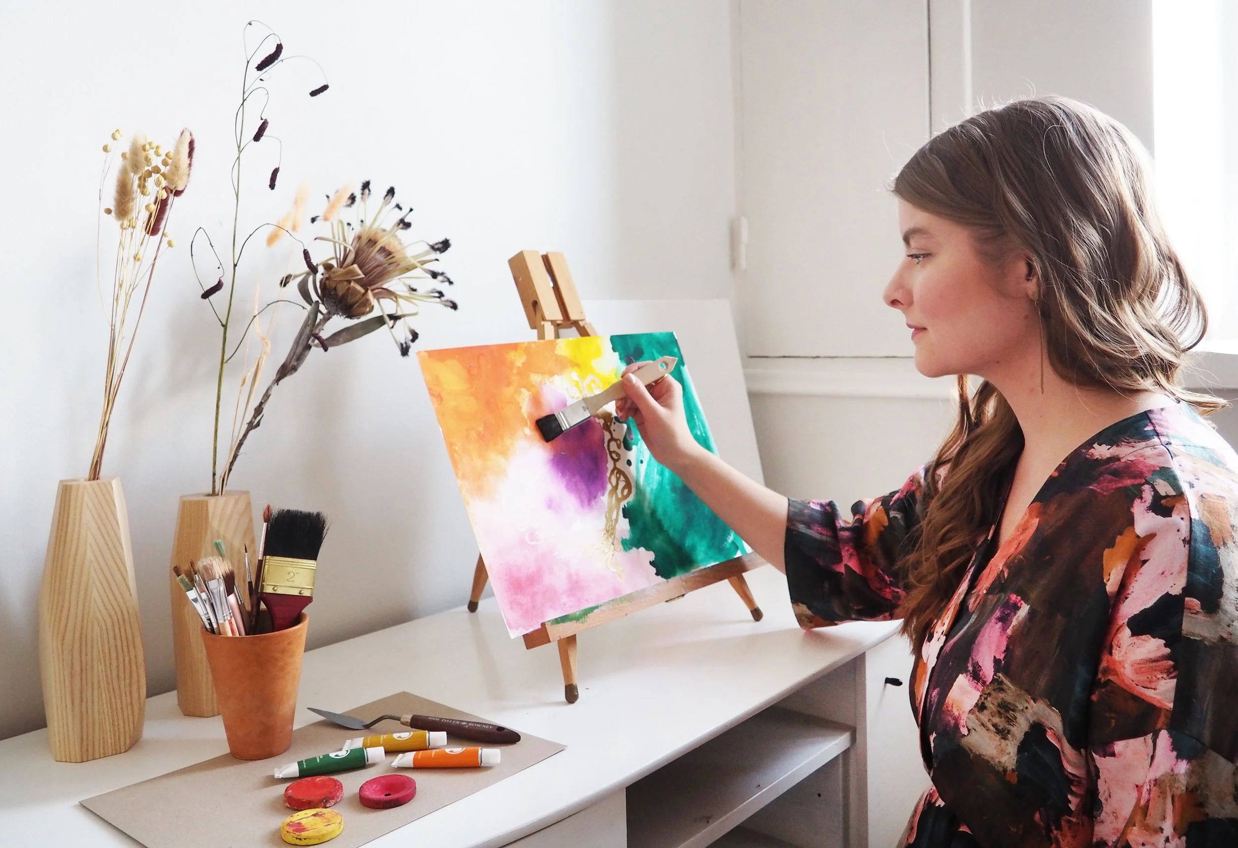 A woman painting with oil paints on a small canvas on a wooden easel, sitting at a white desk in a room with natural light, surrounded by art supplies and dried flowers in vases.