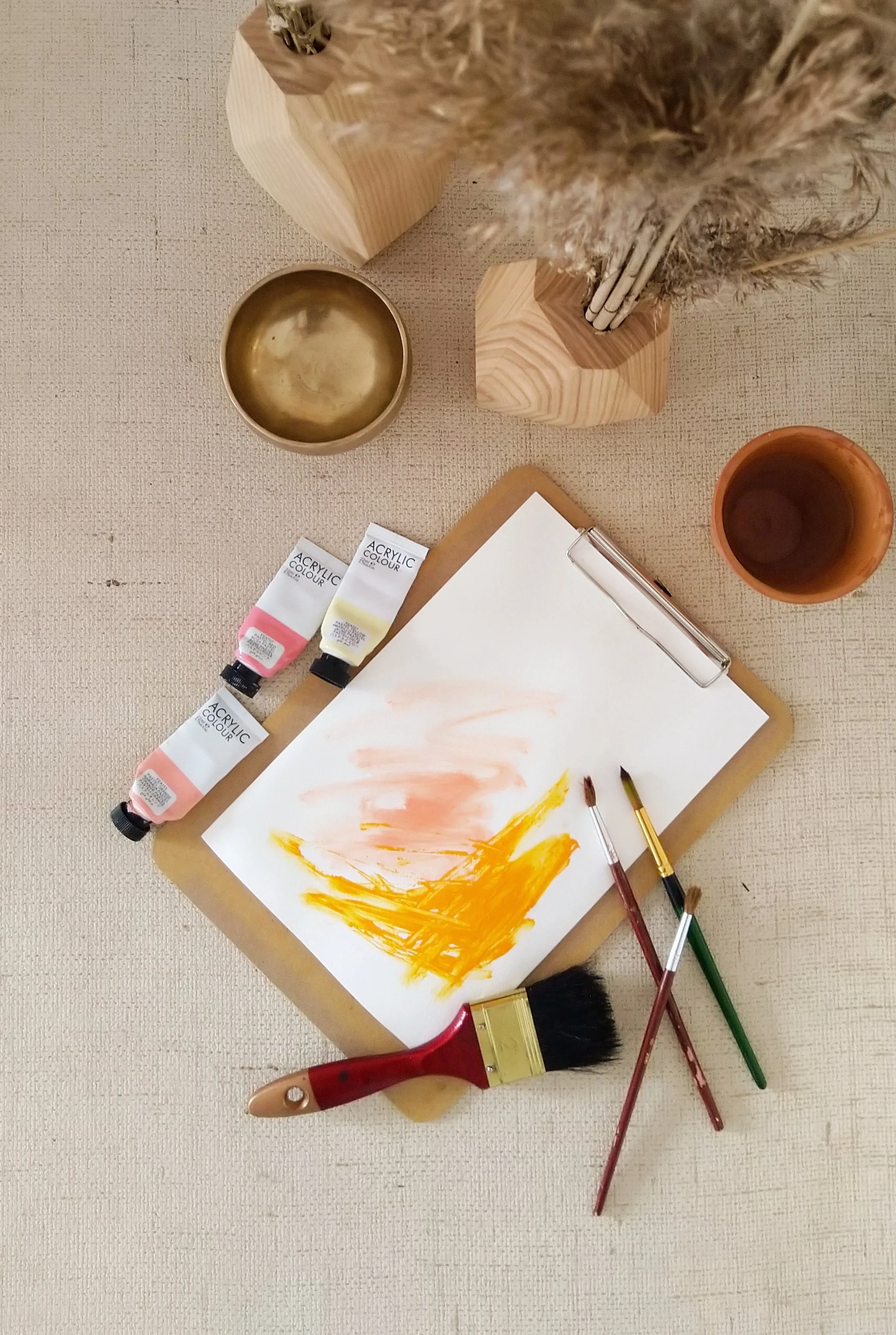 Artist's workspace with a wooden clipboard holding a watercolor painting, three bottles of acrylic paint, three paintbrushes, a large paintbrush, a gold-colored cup, a small copper-colored cup, and two wooden vases containing dried plants, all on a beige textured surface.
