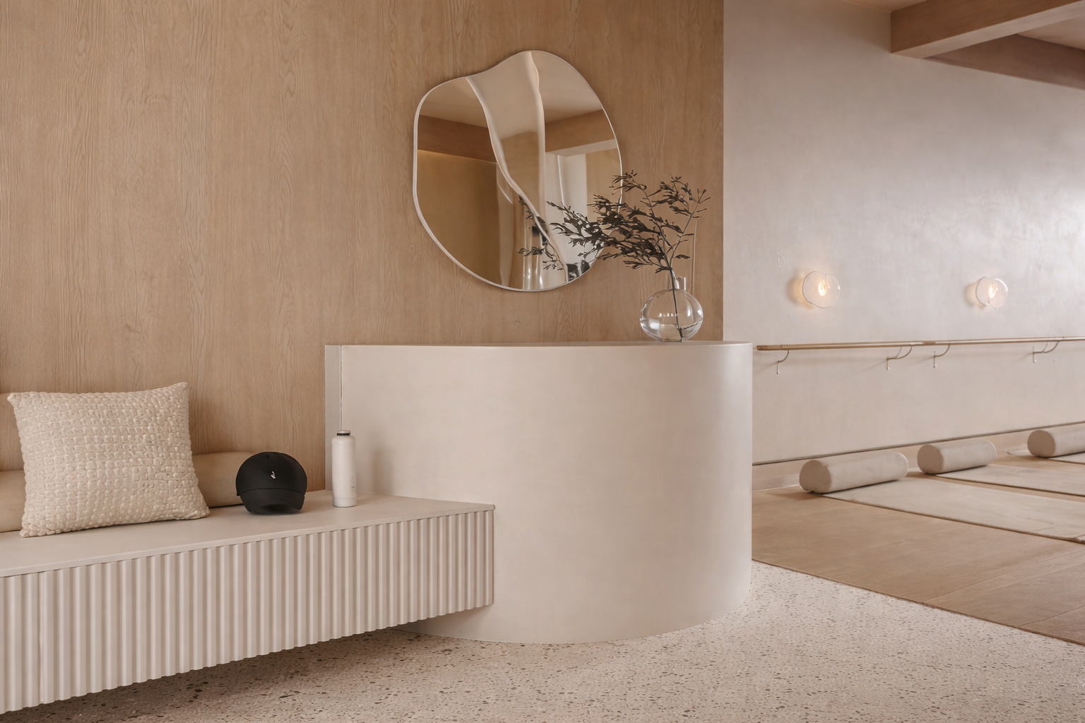 Minimalist interior with a curved white reception desk, a floating bench with a pillow, a black helmet, a white water bottle, and a bowl with plant in a transparent vase. Soft lighting and natural wood elements.