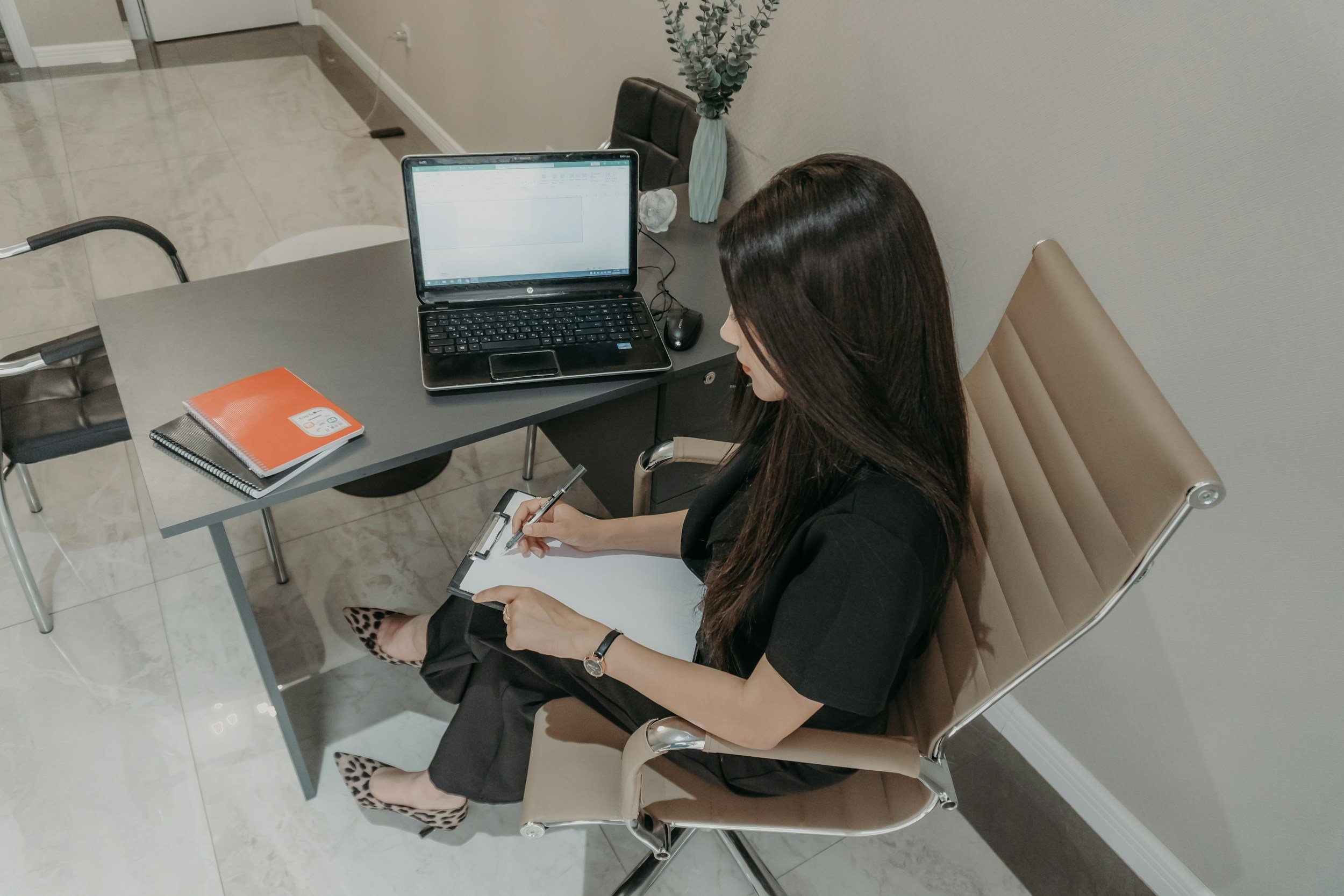 A woman is sitting at a desk taking notes with a clipboard and pen. The desk has a laptop, a red notebook, a black notebook, and a computer mouse. There is a potted plant on the desk and a chair in the background.