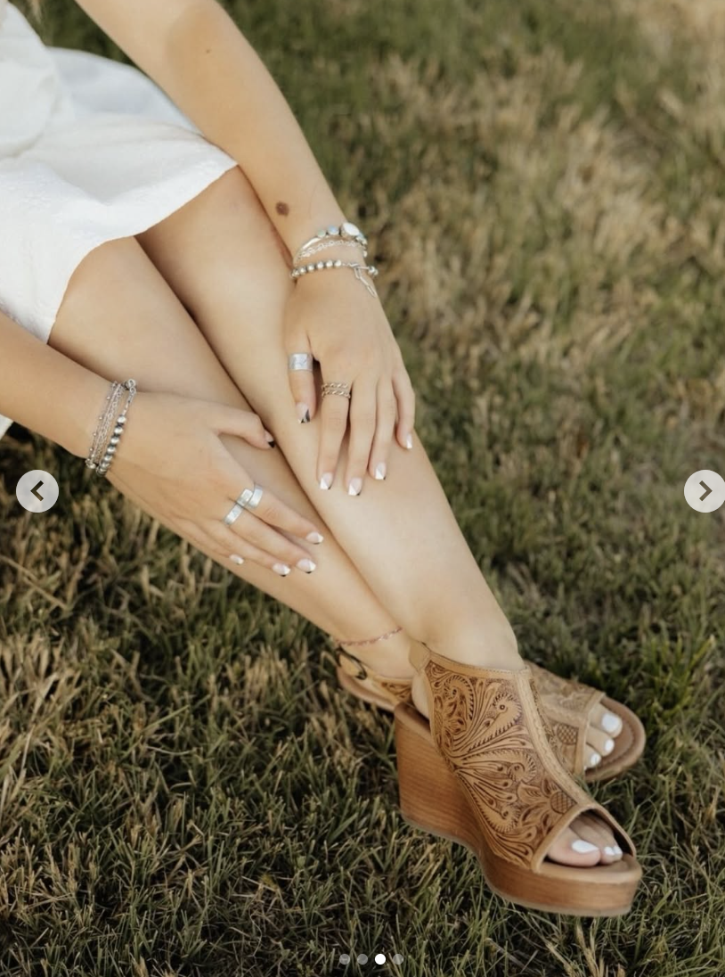 Person sitting on grass, wearing tan open-toe wooden-heeled sandals, white dress, and multiple silver rings and bracelets, with hands resting on their knee.
