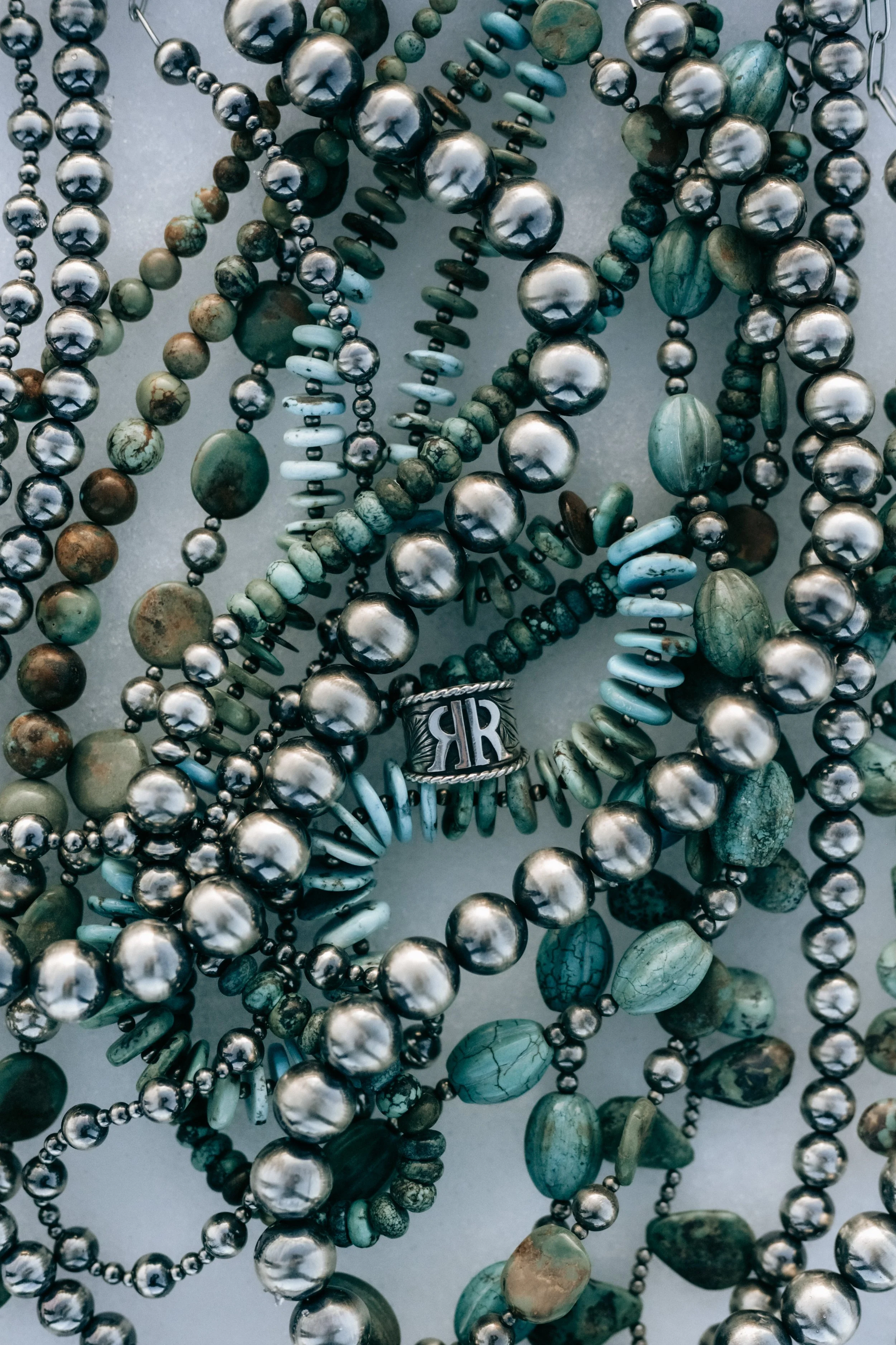 A collection of assorted beaded necklaces, primarily in shades of green and silver, with a silver ring featuring the monogram 'RR' placed among them.