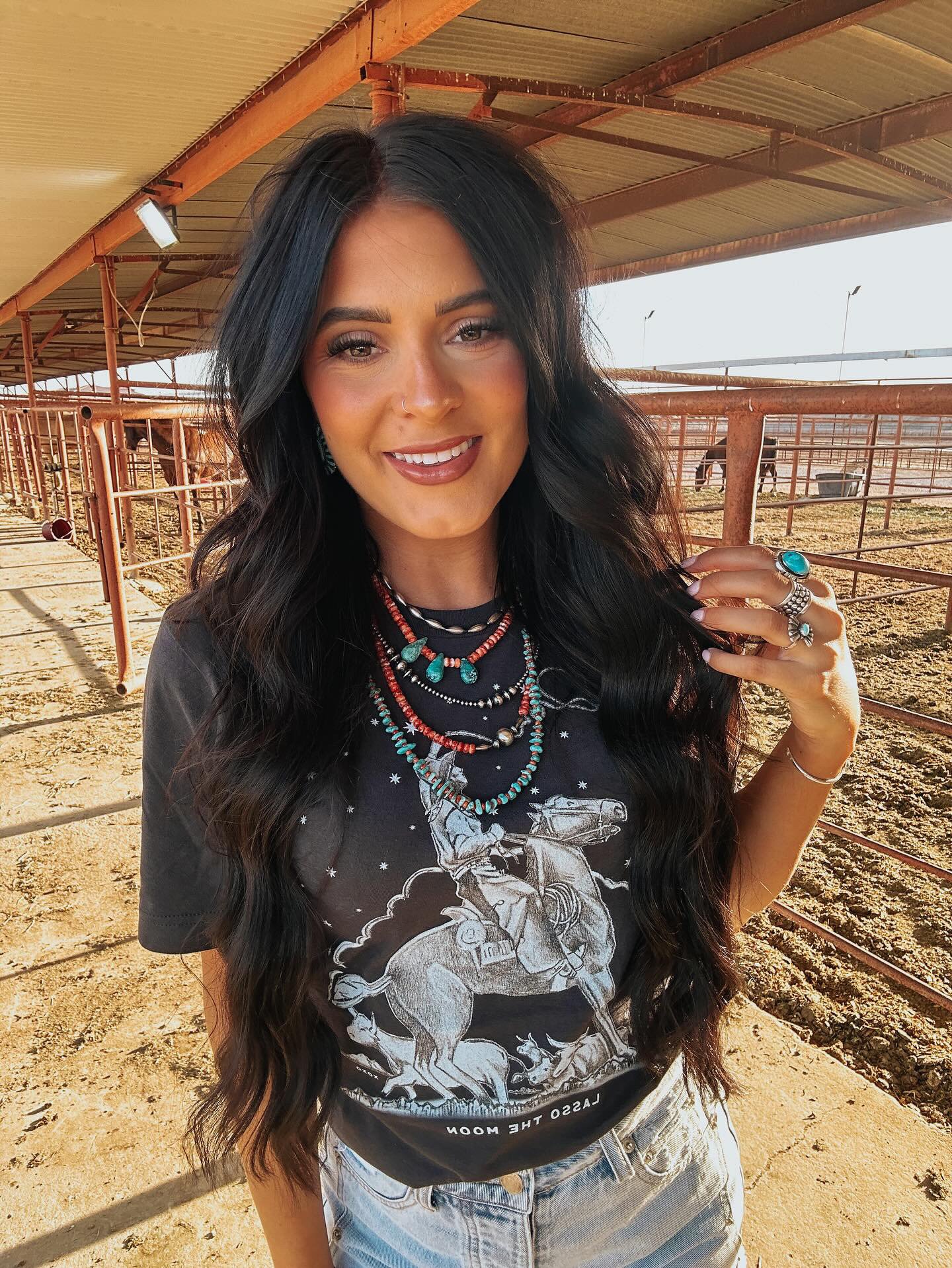 Young woman with long black wavy hair wearing multiple necklaces, rings, a nose ring, and a graphic T-shirt, standing in a livestock pen or barn with metal fencing and a metal roof.