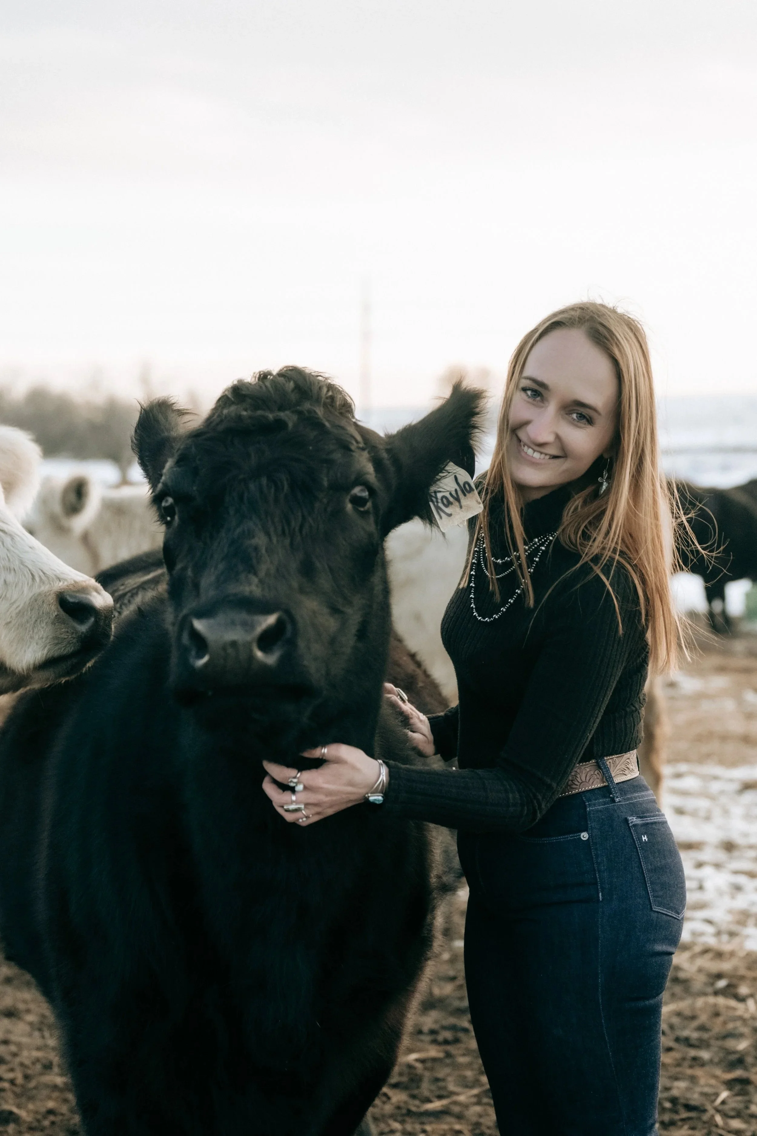 A woman with red hair smiling while holding a black calf on a farm with other cattle in the background.