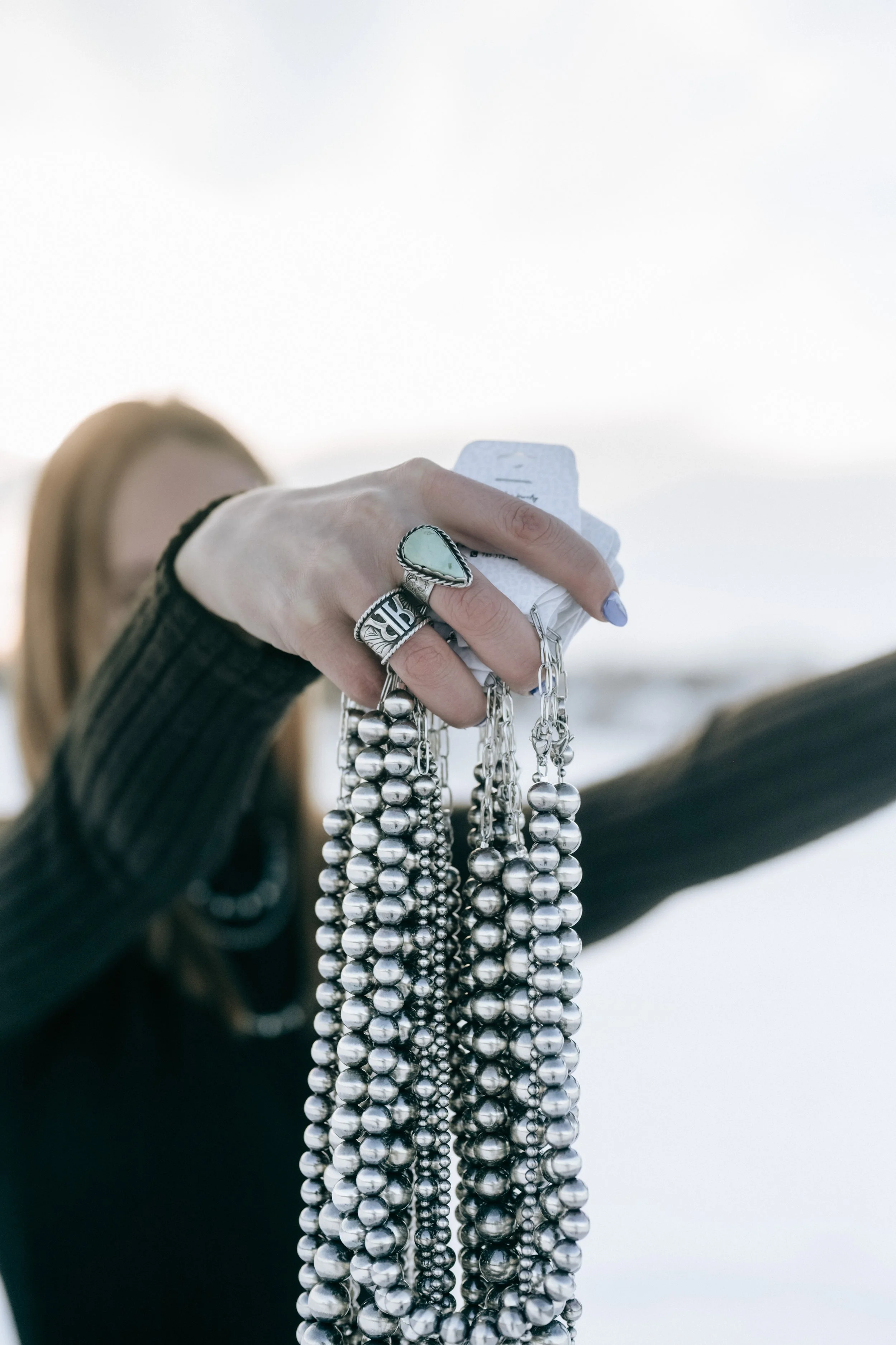 A person's hand holding multiple silver jewelry chains and rings, with a blurred background and part of their arm in a black sleeve.