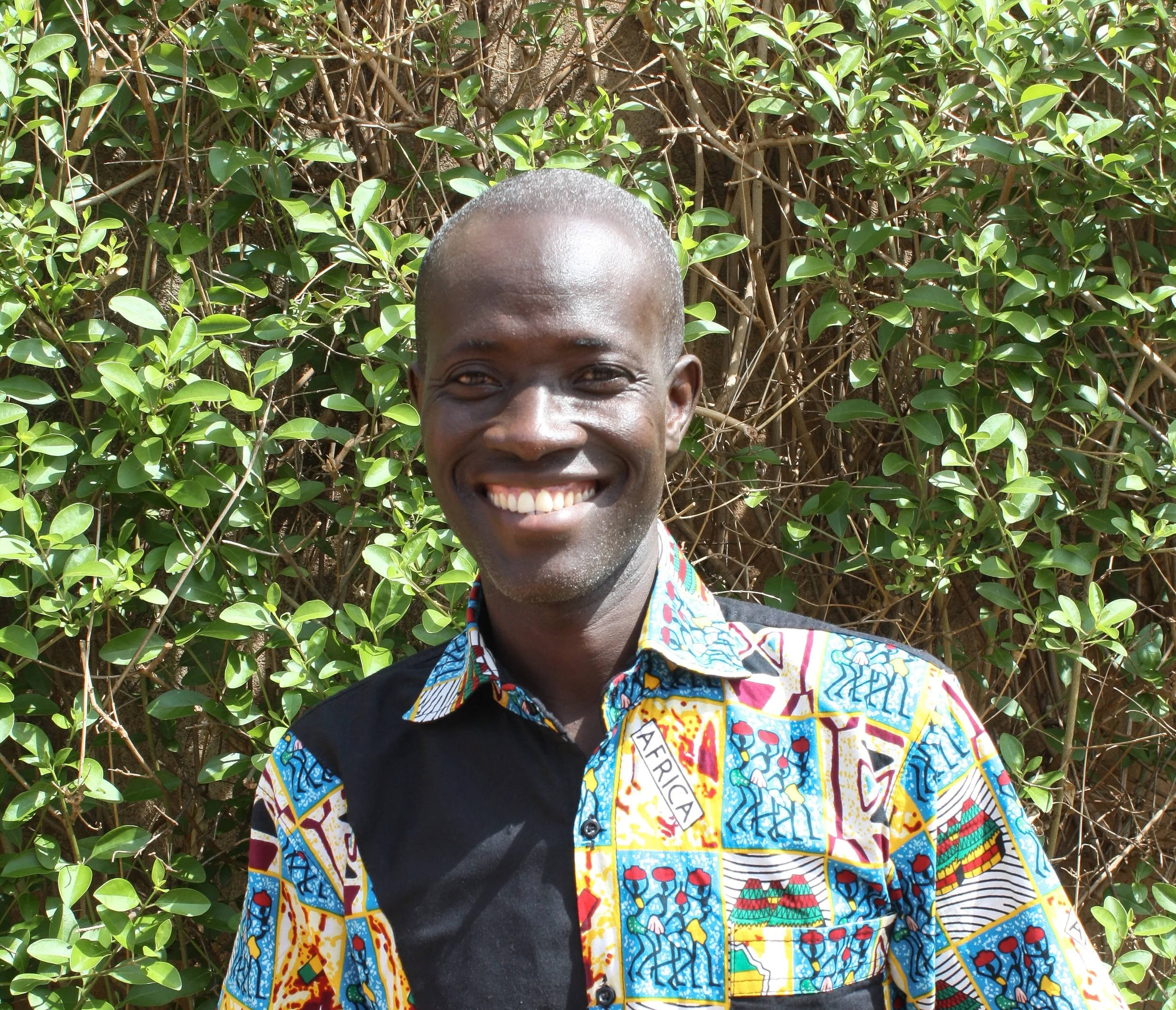 A smiling man with short hair standing in front of a leafy shrub, wearing a colorful African print shirt with the label 'AFRICA' on it.