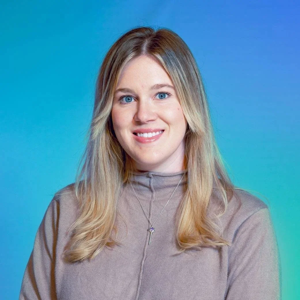 A woman with long blonde hair, blue eyes, smiling, wearing a beige turtleneck top and a silver necklace with a key-shaped pendant, standing against a blue-green gradient background.