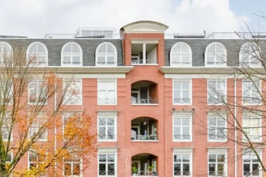 Bovenaan van een modern gebouw met meerdere ramen en een gevel met rood en wit baksteen, omringd door herfstbomen.