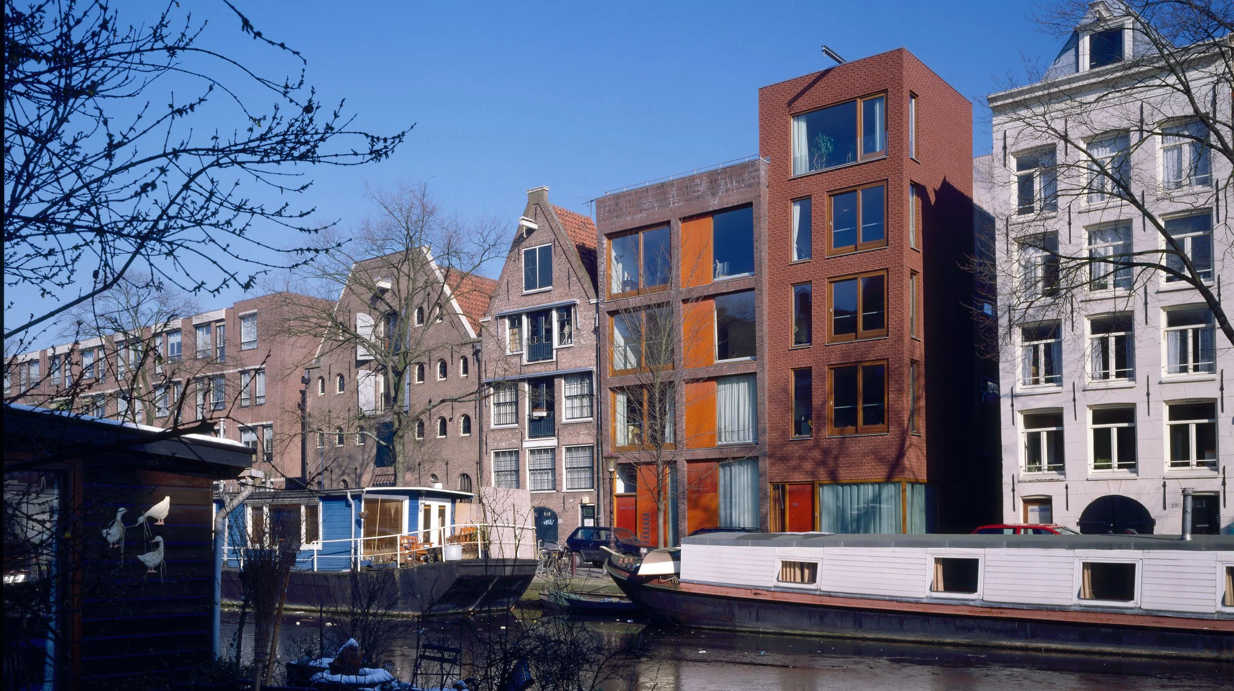 Een kanaal met twee boten, omringd door gebouwen inclusief een modern rood-bruin gebouw met grote ramen en traditionele huizen, onder een blauwe hemel.