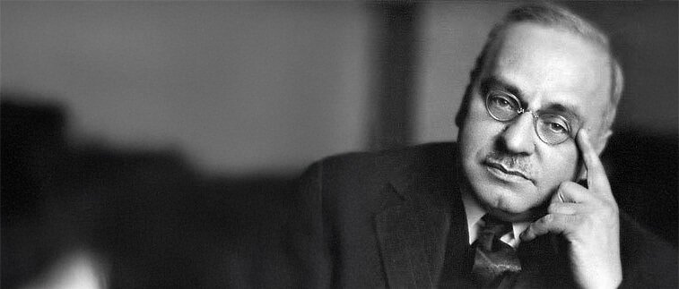 Foto von Alfred Adler. Schwarz-weiß Foto eines Mannes mit Brille, welcher Denkerpose macht, das Kinn mit der Hand stützt, in formeller Kleidung.