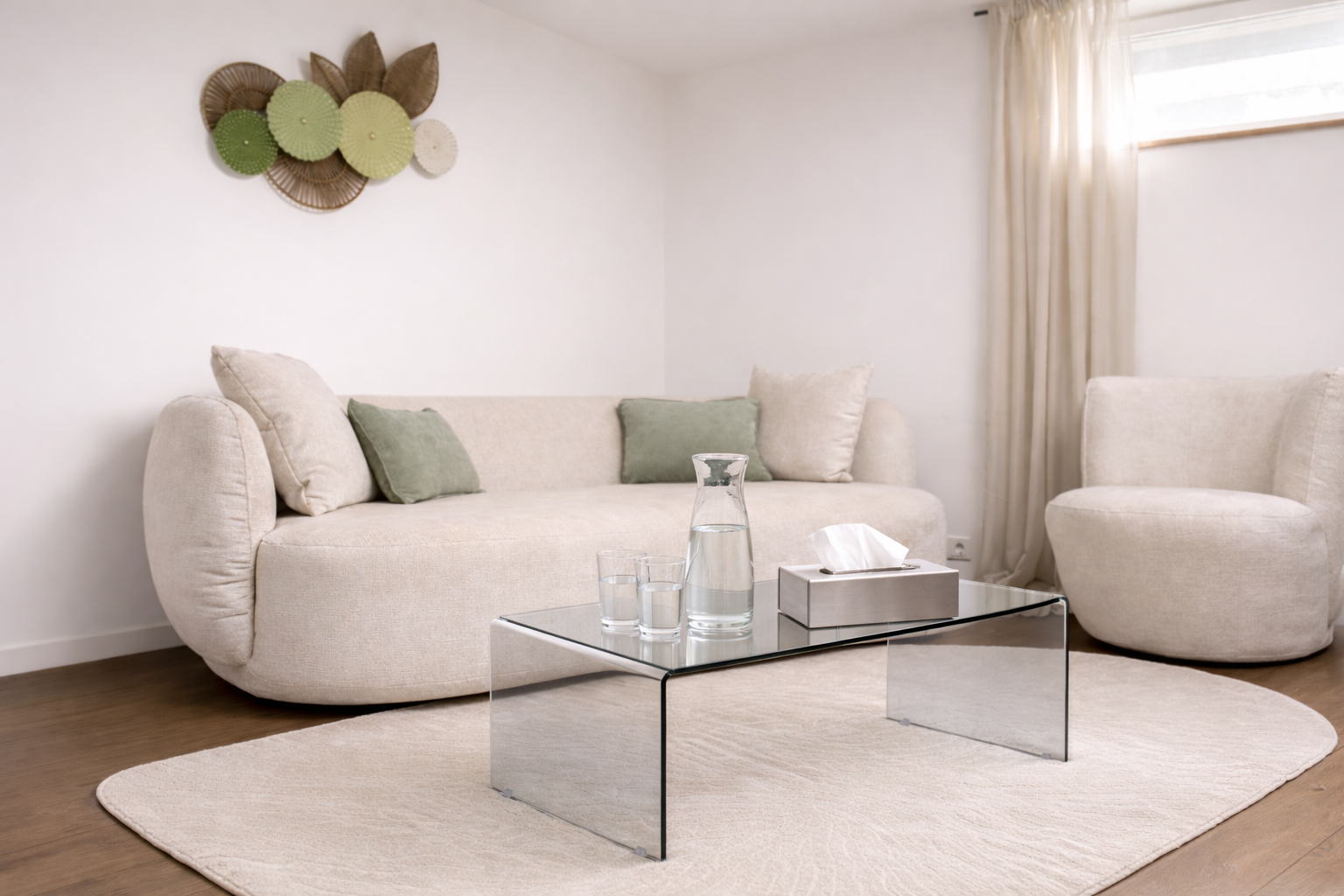Ein helles, modernes Wohnzimmer mit weißen Sofas, einem Glastisch, einem Wasserkrug, zwei Gläsern Wasser, einer Kiste mit Taschentüchern, einem weißen Teppich und Wanddekorationen aus geflochtenen Körben und Stoffblumen.