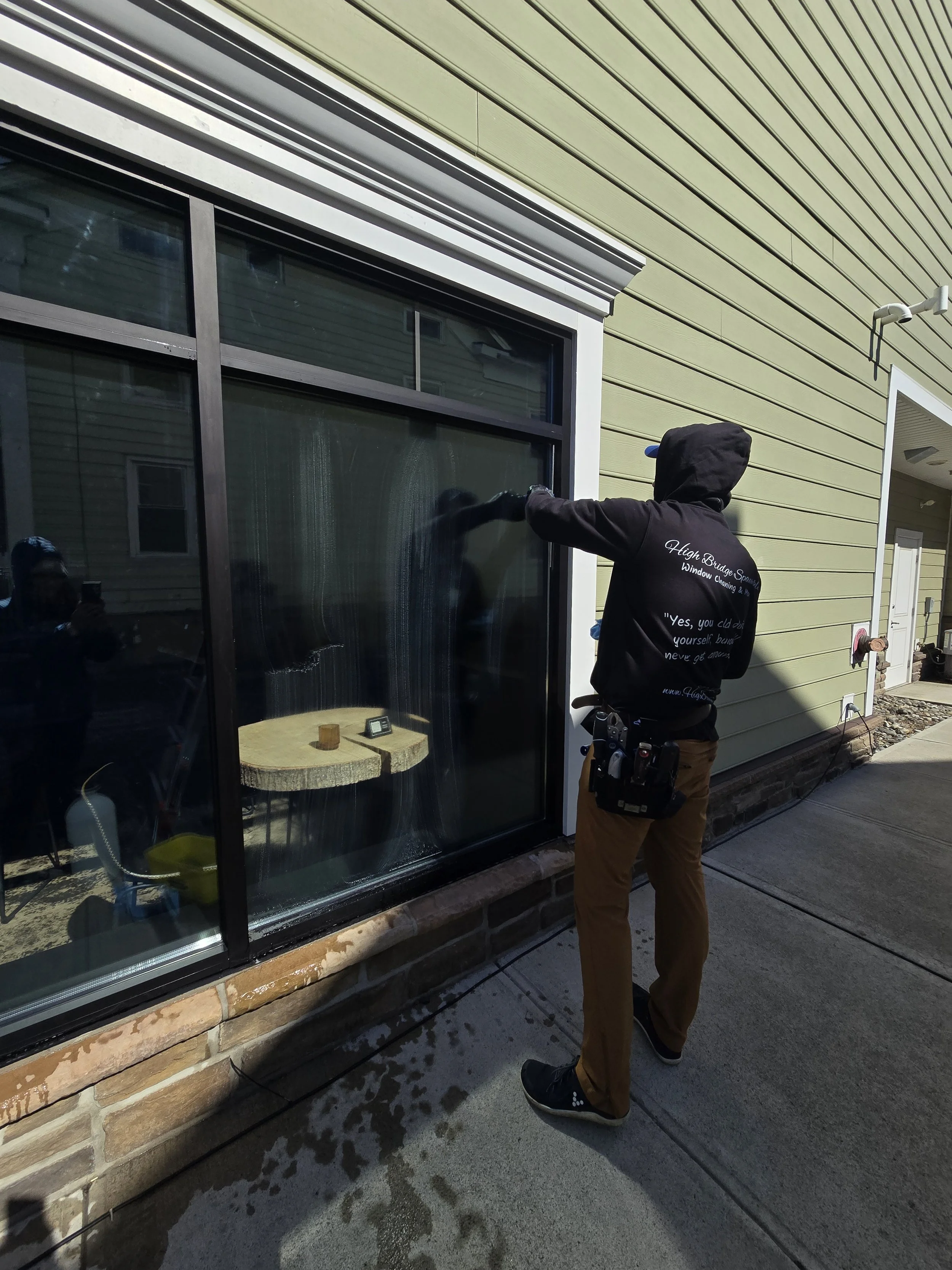 A worker cleaning the outside glass of a large window on a yellow house using a squeegee on a sunny day.