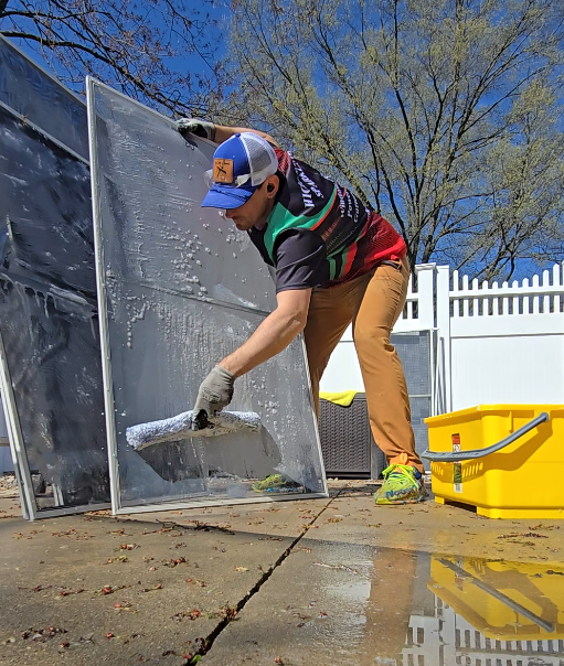 A person cleaning a large glass window outdoors on a sunny day, with a yellow bucket nearby and a white picket fence in the background.