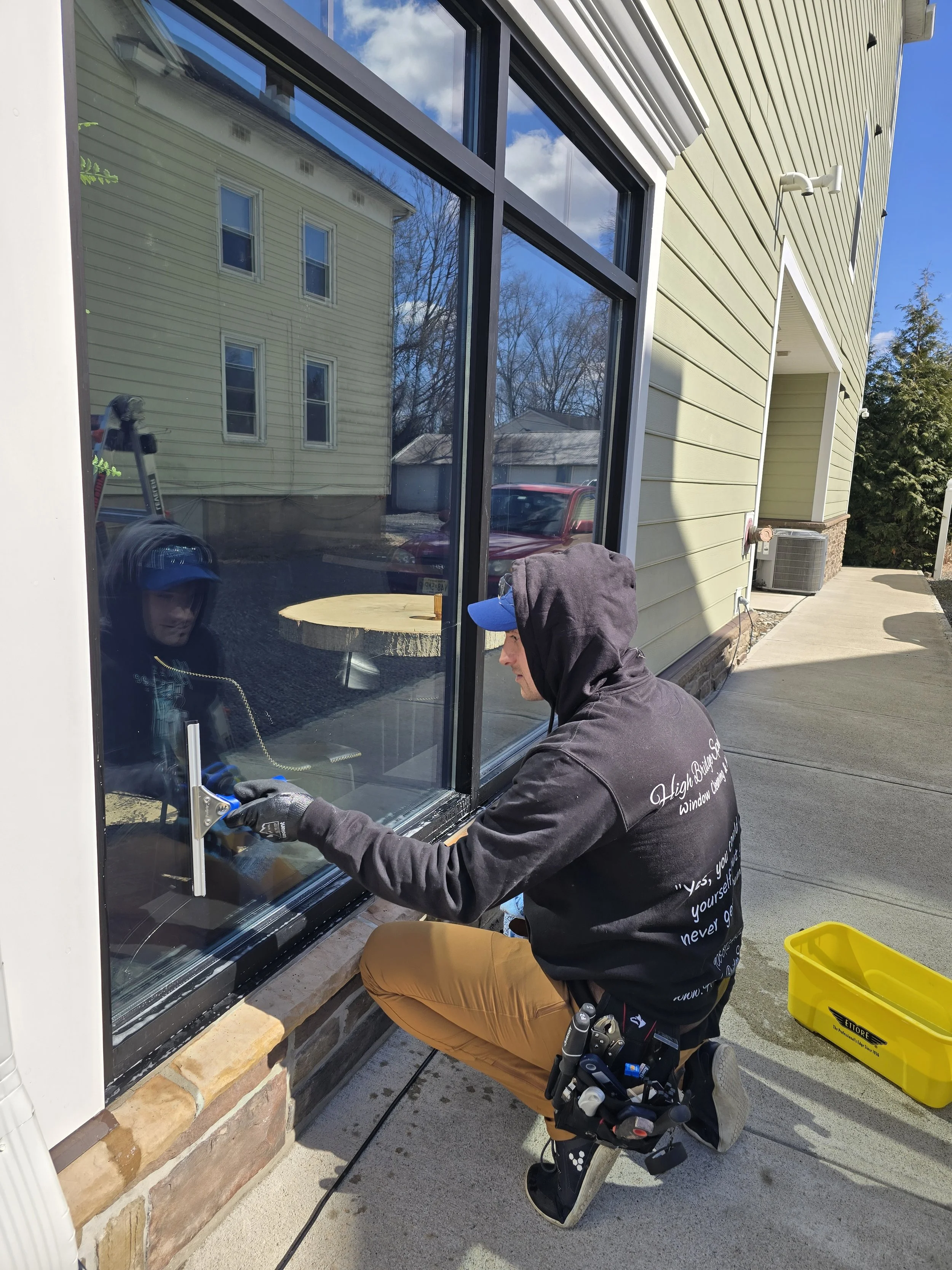 A worker installing or repairing a large window on a house exterior, wearing a black hoodie, brown pants, and kneeling on the ground with tools attached to his belt while working outside on a sunny day.