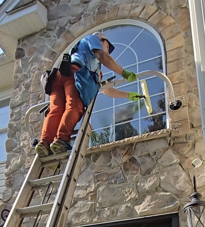 A person on a ladder cleaning a large arched window on a stone house using a squeegee, wearing a hat and green gloves.