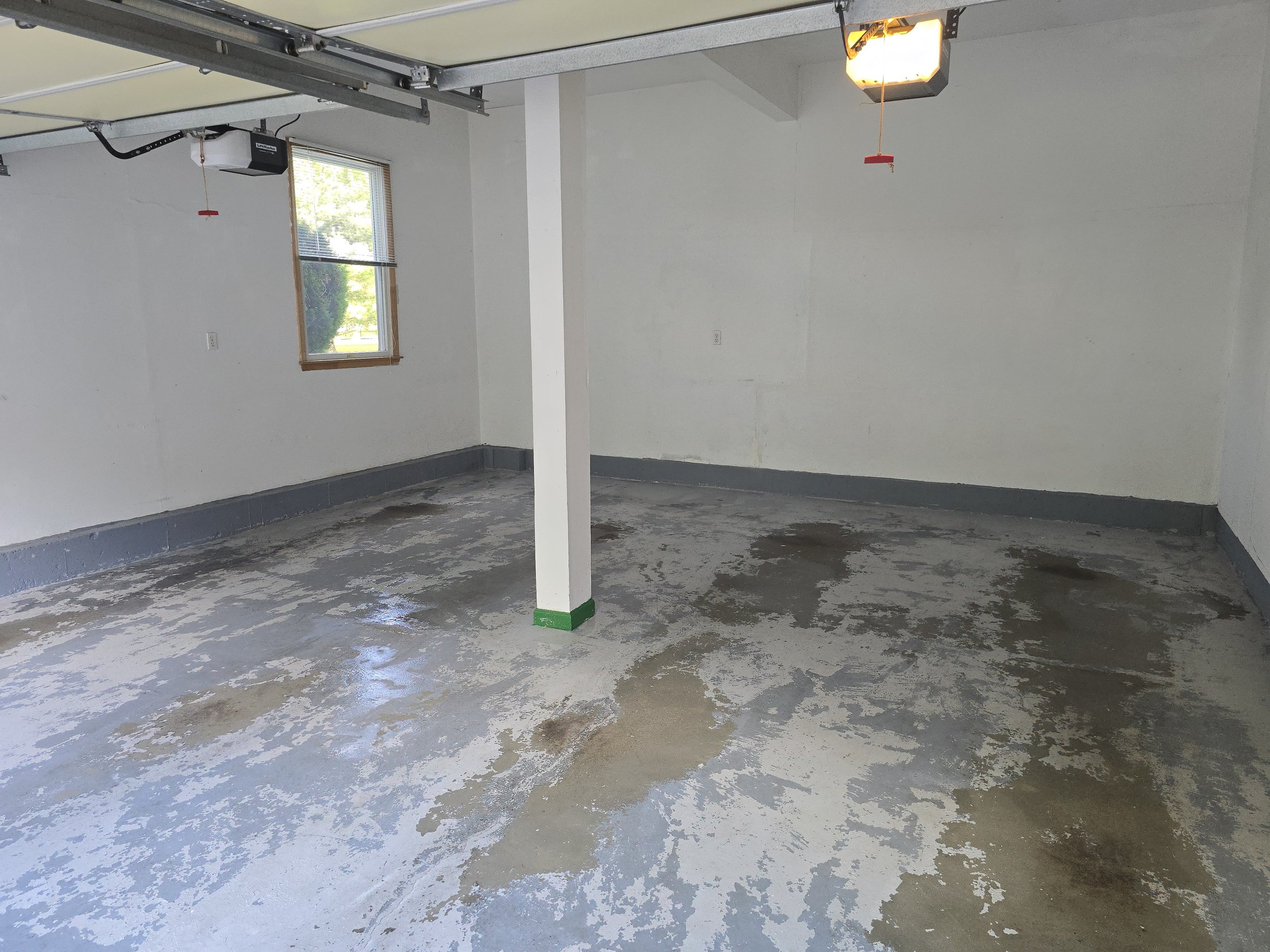 Empty garage with concrete floor, white walls, window, overhead garage door opener, and support pillar with green tape at its base.