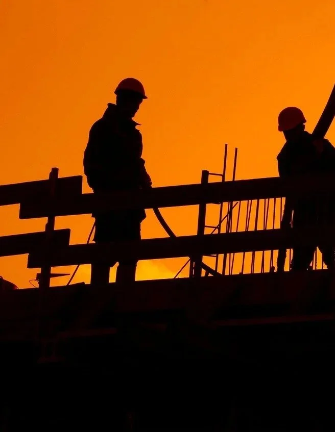 Silhouettes of two construction workers wearing helmets on a building site during sunset or sunrise.